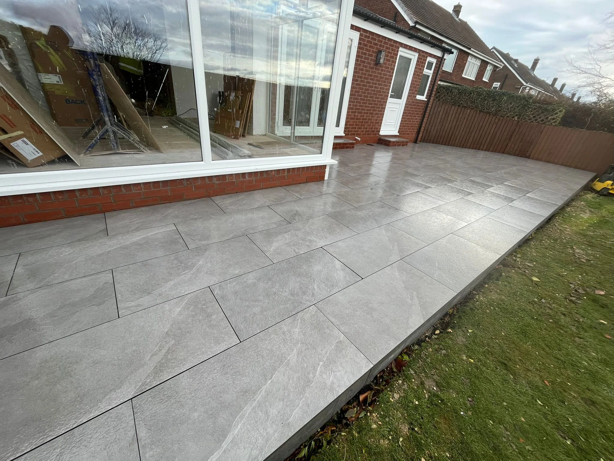 Newly installed gray tiled patio outside a house with a brick wall and glass conservatory, with some construction materials inside visible through the window.
