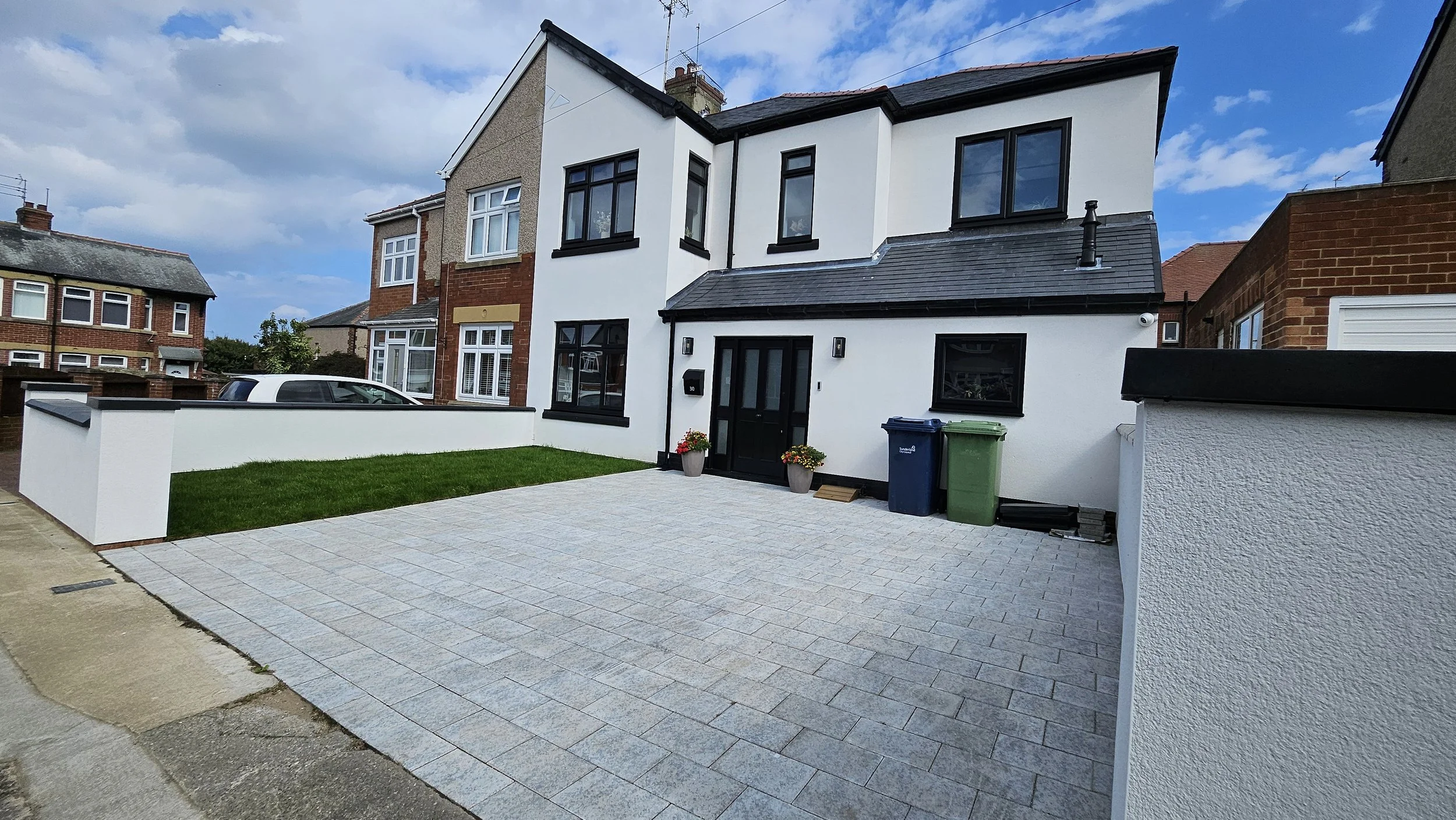 Front view of a modern two-story house with white exterior walls, black window frames, and a darker roof. The house has a paved driveway, two potted plants near the front door, and a small strip of grass. The sky is partly cloudy.