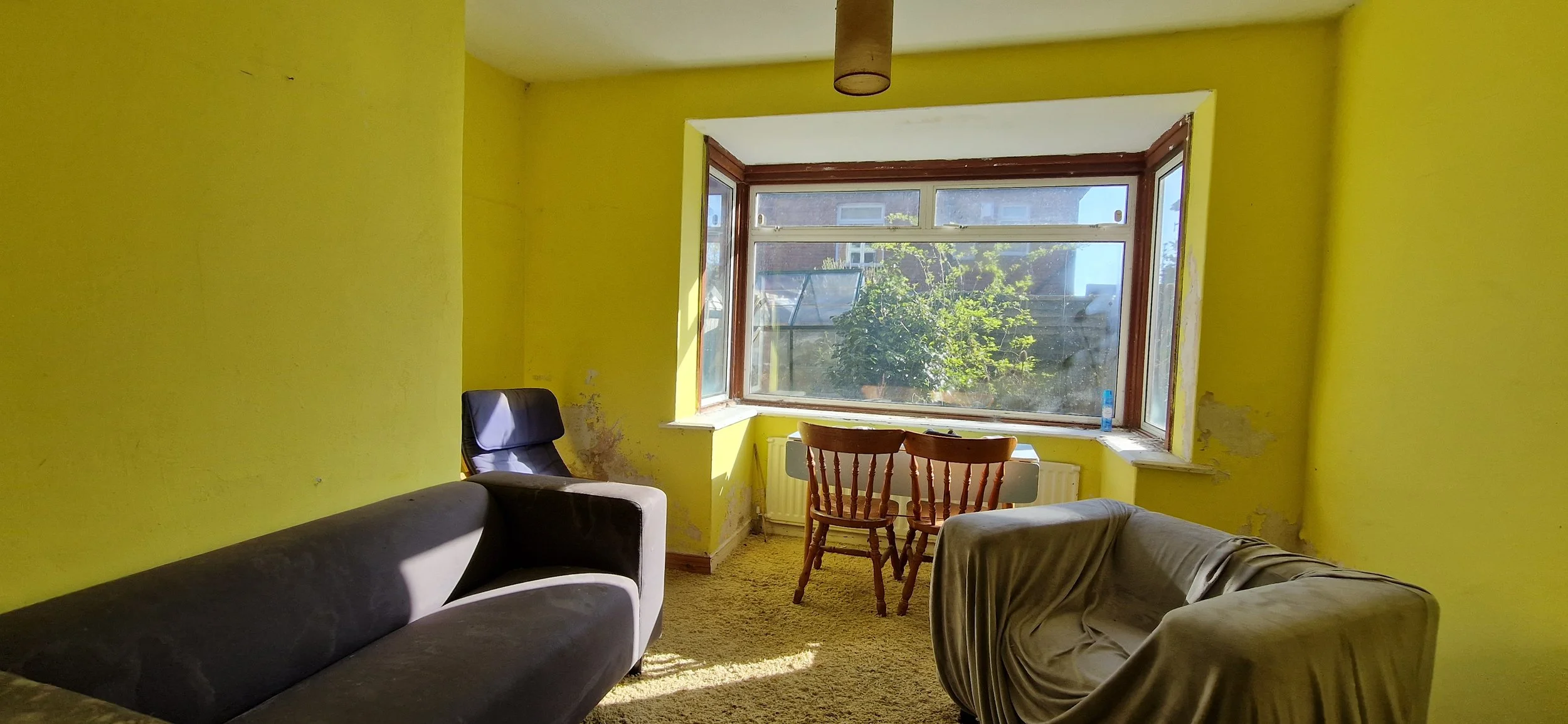Living room with yellow walls, a large bay window, a dark sofa, a chair, a small round table, and a beige armchair with a cloth cover. Sunlight streams through the window, illuminating the room.