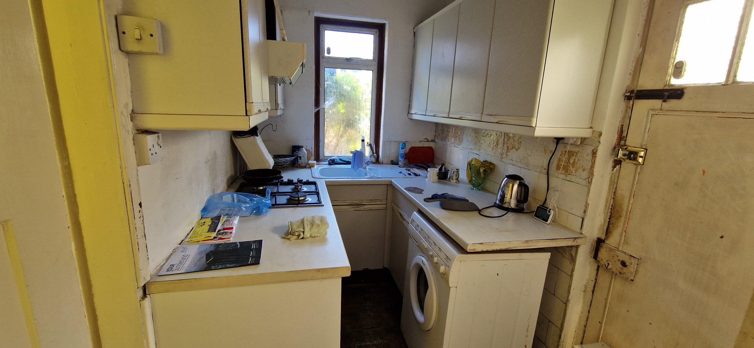 Small, cluttered kitchen with white cabinets, a washing machine, a gas stove, and various kitchen items on the countertops, including a kettle, phone, and paper towels, with a window overlooking greenery.