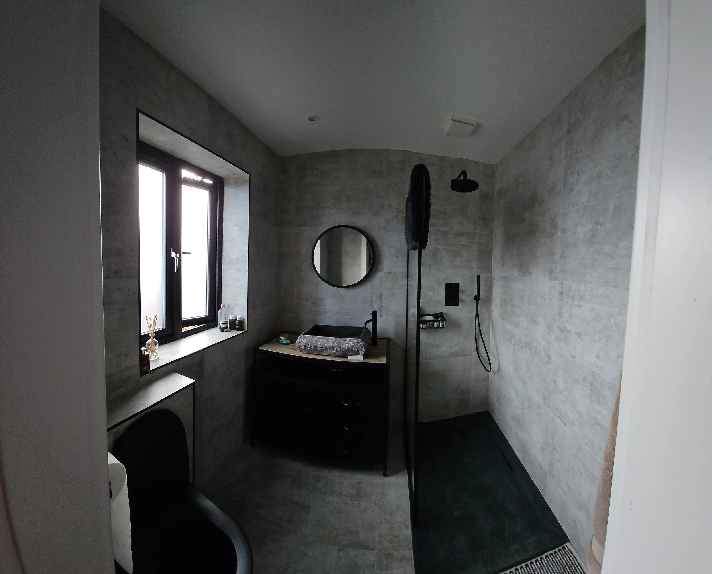 Bathroom with dark window, grey tiled walls, circular mirror, sink with marble basin, black vanity, shower area with rain shower head and handheld shower, black towel or cloth hanging, small toiletries on shelf, black floor mat, and white toilet part