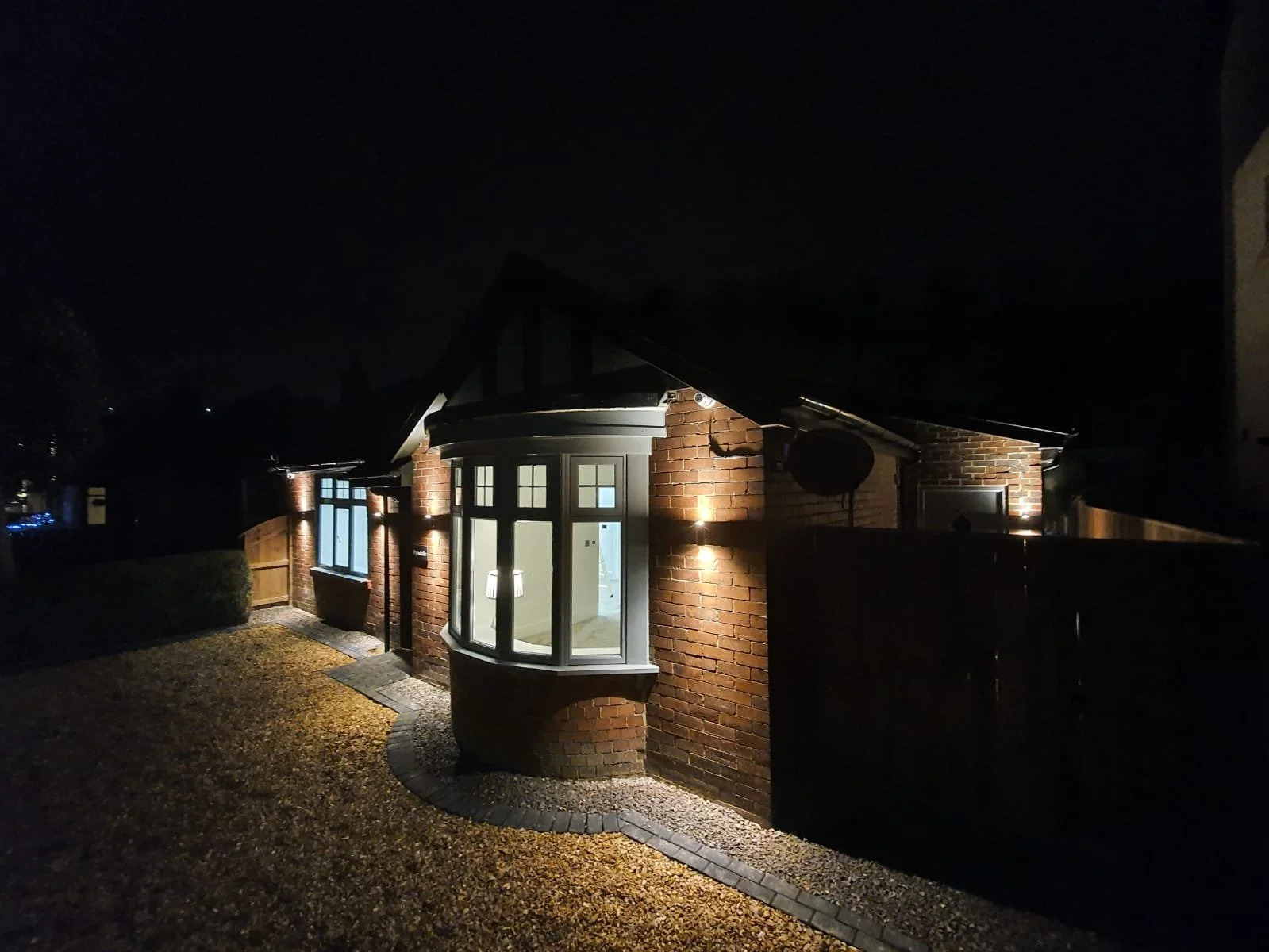 Nighttime view of a house with brick walls, illuminated windows, and exterior lights, surrounded by a fenced yard with gravel pathway.