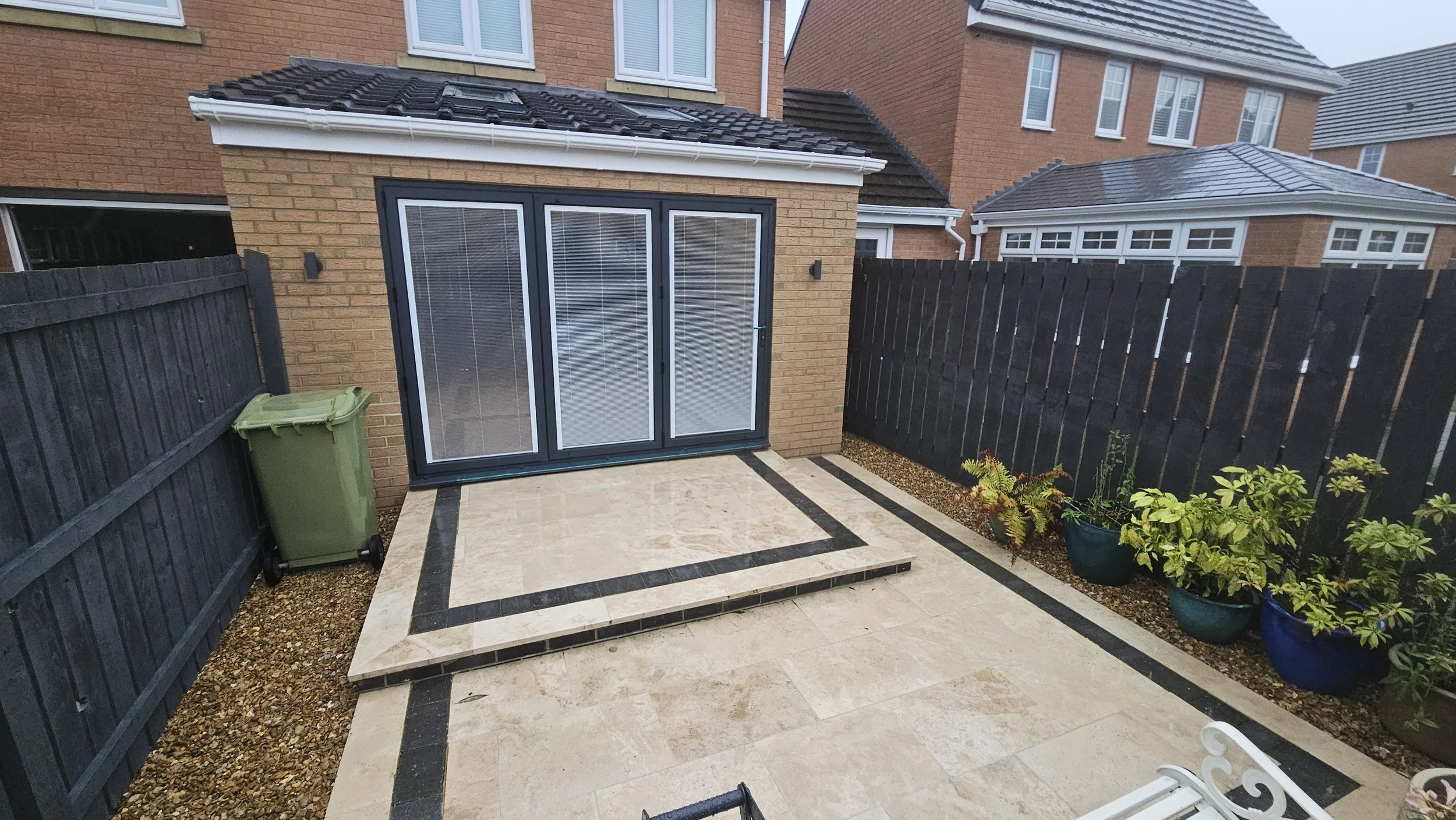 Backyard patio with beige stone tiles and black borders, small set of steps leading to glass patio doors, wooden black fence, potted plants on the right, and a green trash bin on the left.