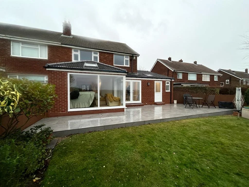 Backyard with grass lawn, stone patio, outdoor furniture, and large house with a brick exterior and a conservatory.