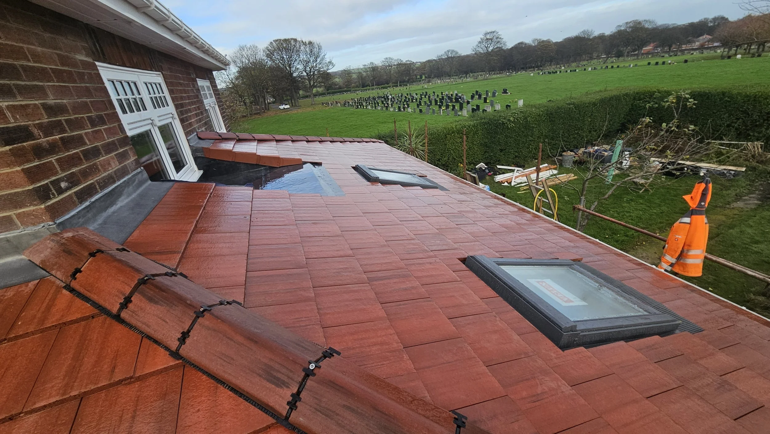 View from a house rooftop showing new red tiled roof, skylights, and a worker in orange safety gear on the scaffolding, with a grassy cemetery and trees in the background.
