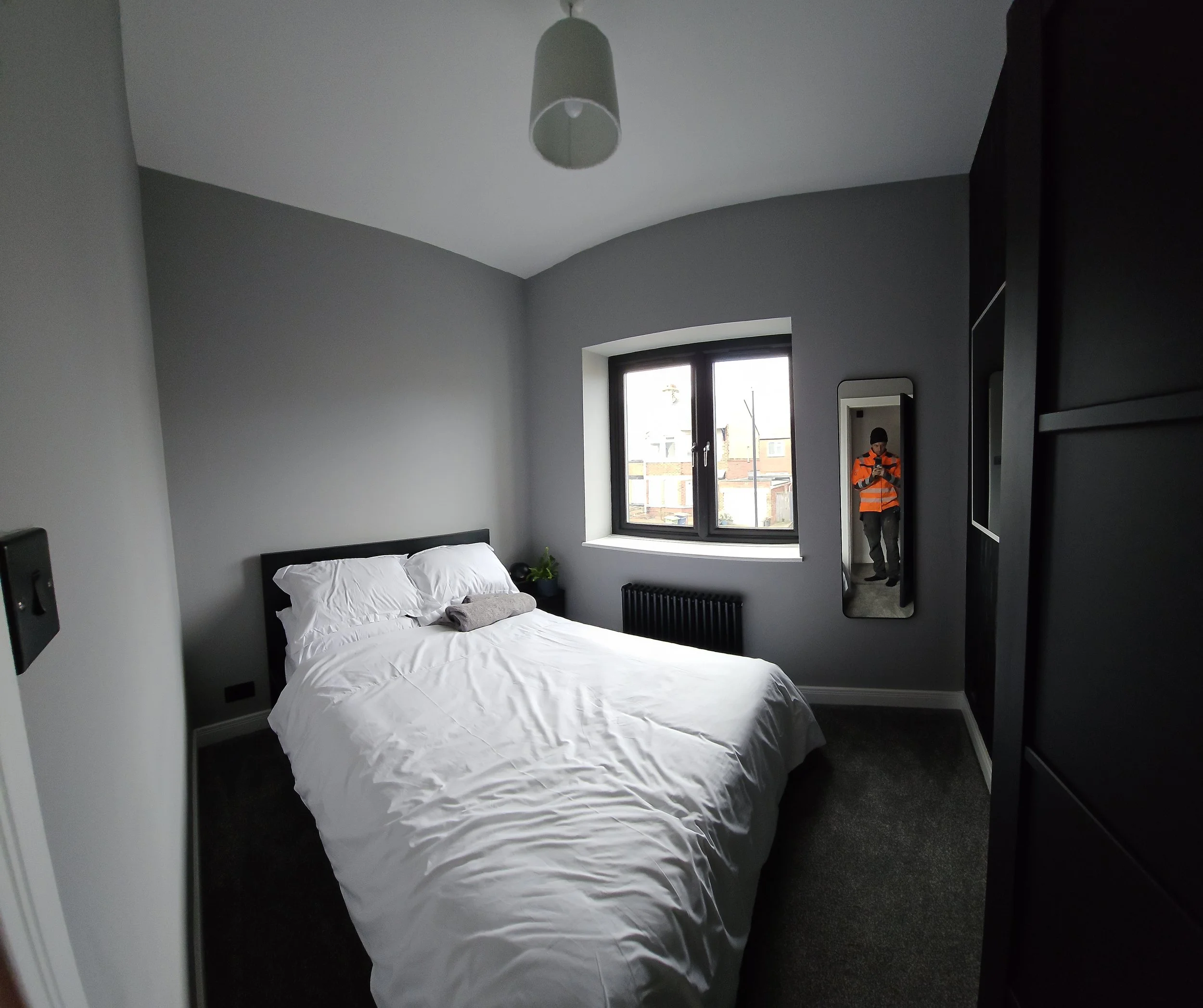 A bedroom with gray walls, a window showing buildings outside, a bed with white bedding and pillows, a small side table with a plant, a floor-length mirror, and a person wearing an orange safety vest taking a photo reflected in the mirror.