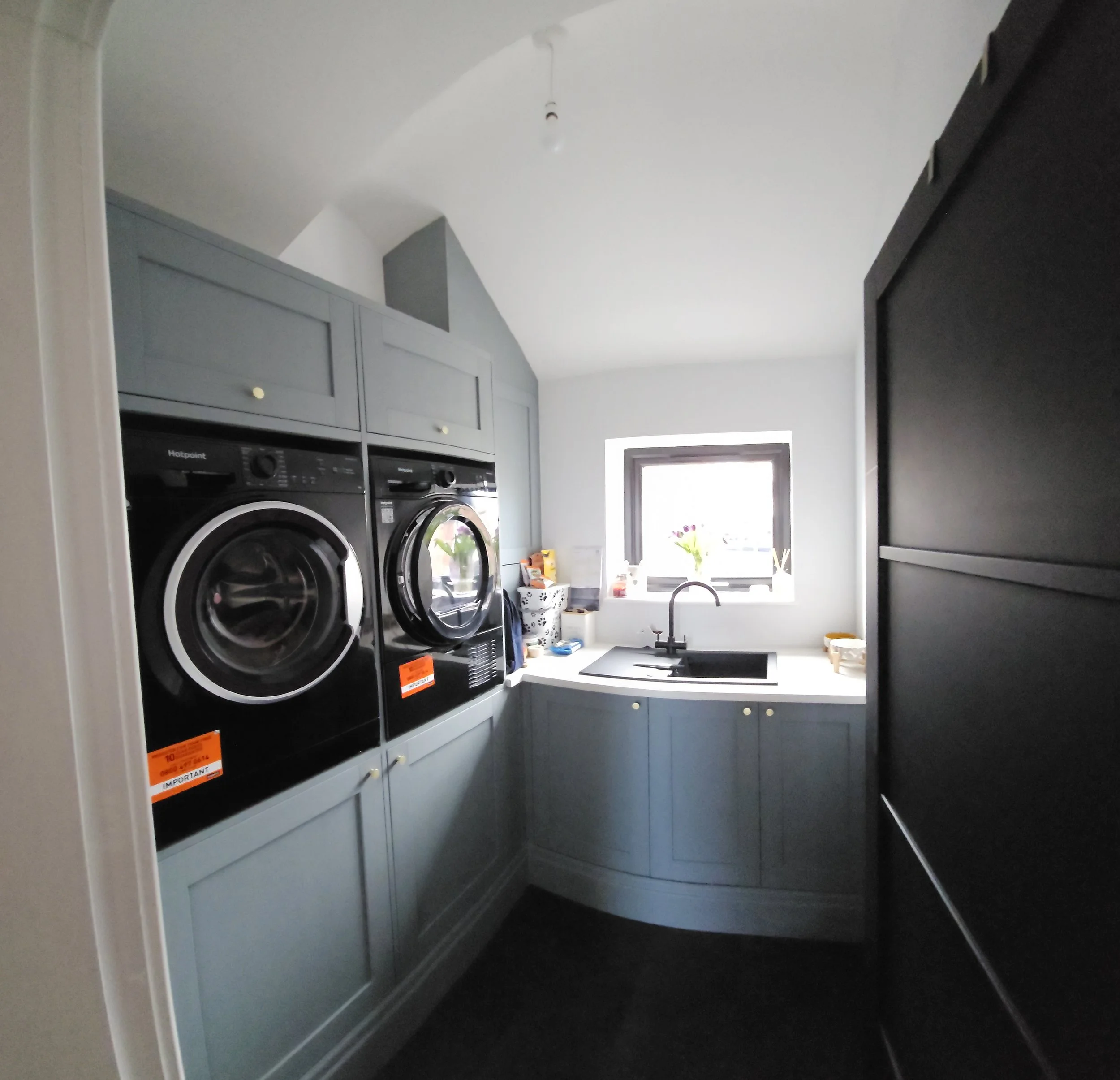 Laundry room with two front-loading black washing machines, gray cabinetry, a small sink, a window with a flower vase, and a black refrigerator on the right side.
