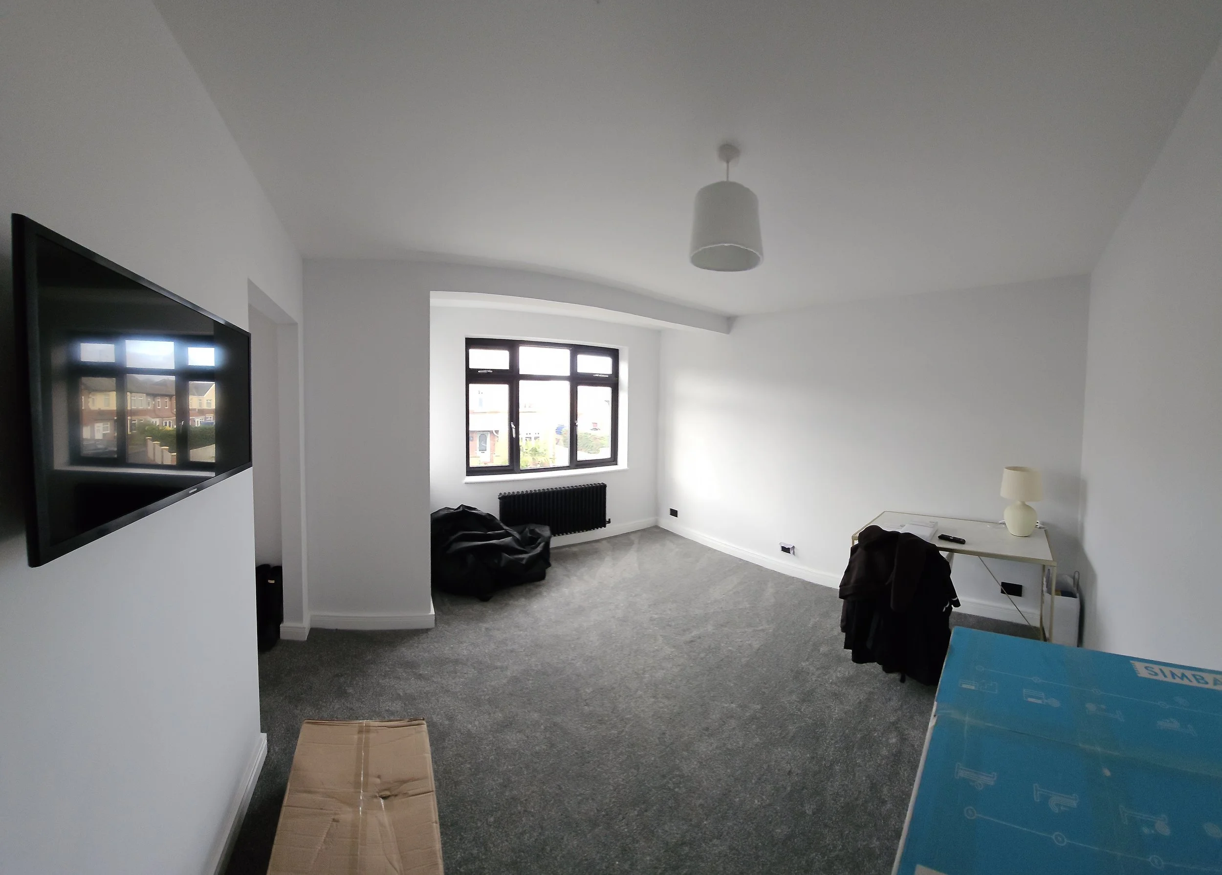 An empty room with white walls and a gray carpeted floor. A window at the far end of the room lets in natural light. A flat-screen TV is mounted on the left wall. There is a white table with a lamp on it and a black jacket on the right side. A cardbo