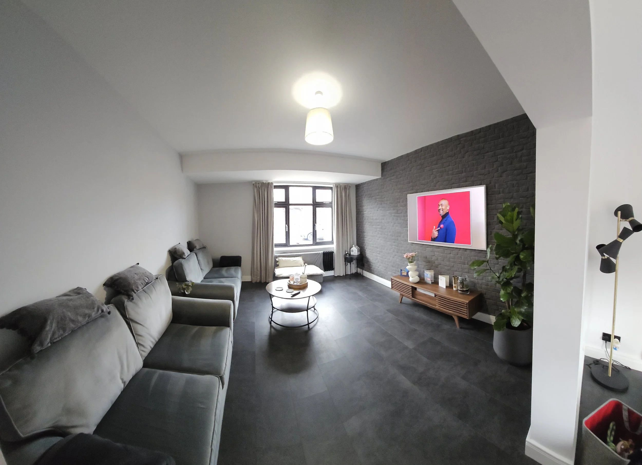A modern living room featuring three grey sofas with cushions, a round coffee table, a TV mounted on a dark brick accent wall, a wooden console below the TV, a potted plant, a small side table near the window, window with beige curtains, and a standi