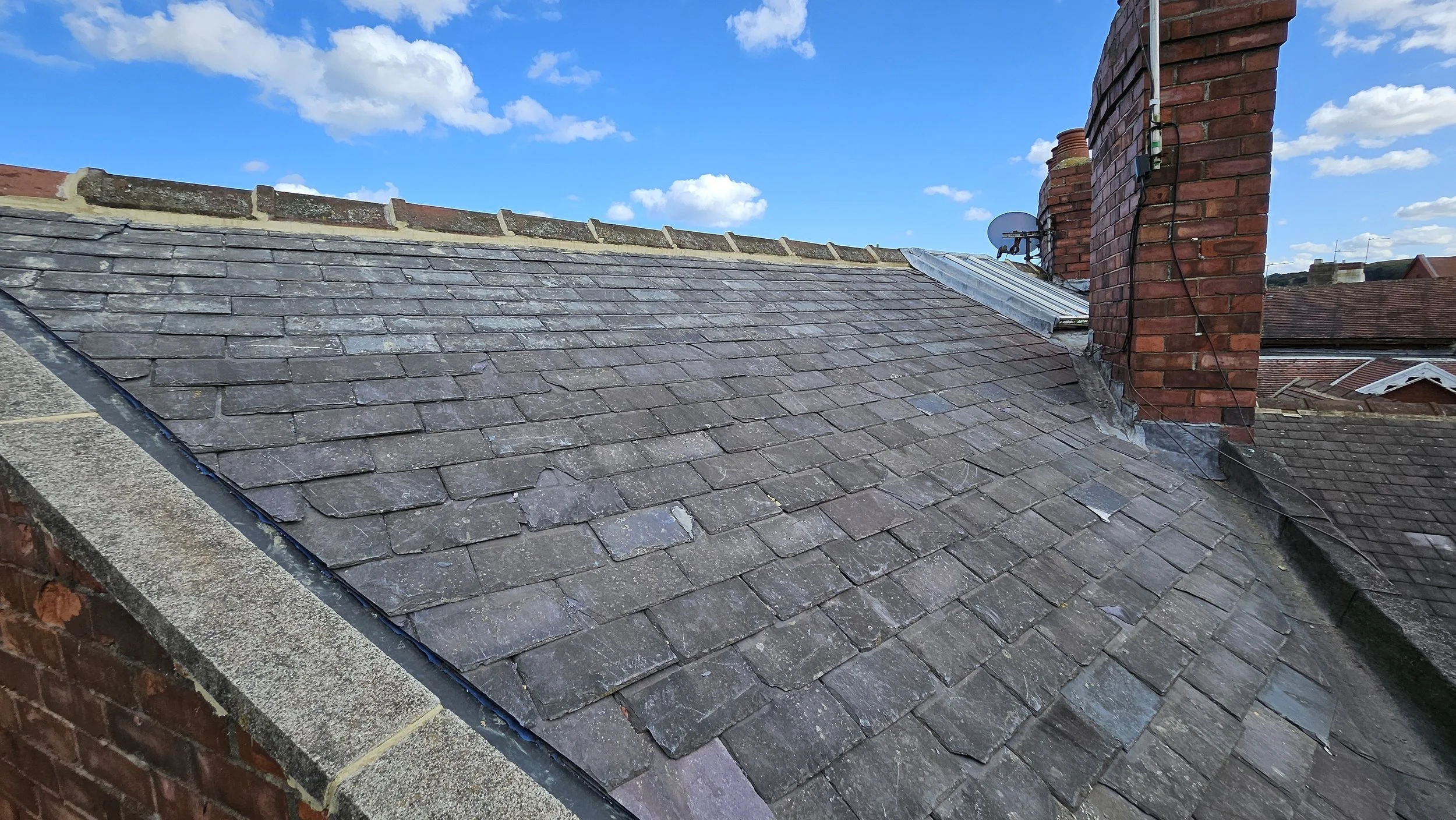 A sloped rooftop with weathered gray shingles, brick chimneys, and a satellite dish under a partly cloudy sky.