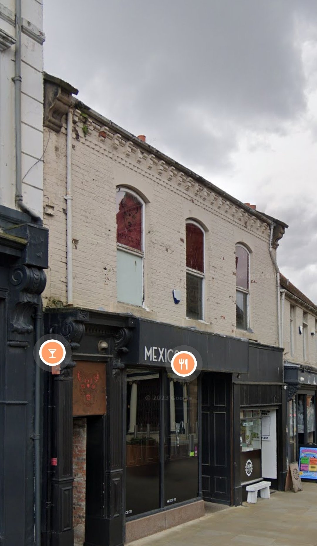 Exterior view of a building with a black storefront that has a sign reading 'MEXICO'. The building has an old brick upper level with three windows and decorative brickwork at the roofline. Overcast sky with dark clouds.