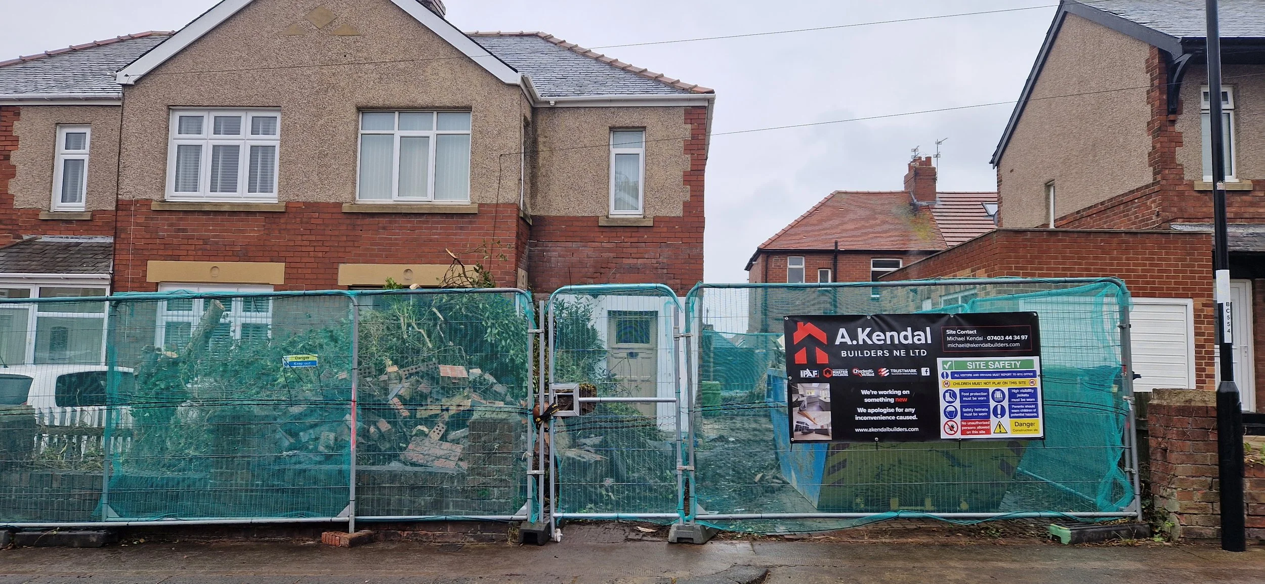 A construction site enclosed by a green safety fence with a sign, showing debris and a fallen tree in front of two brick houses in a neighborhood. The sky is cloudy.