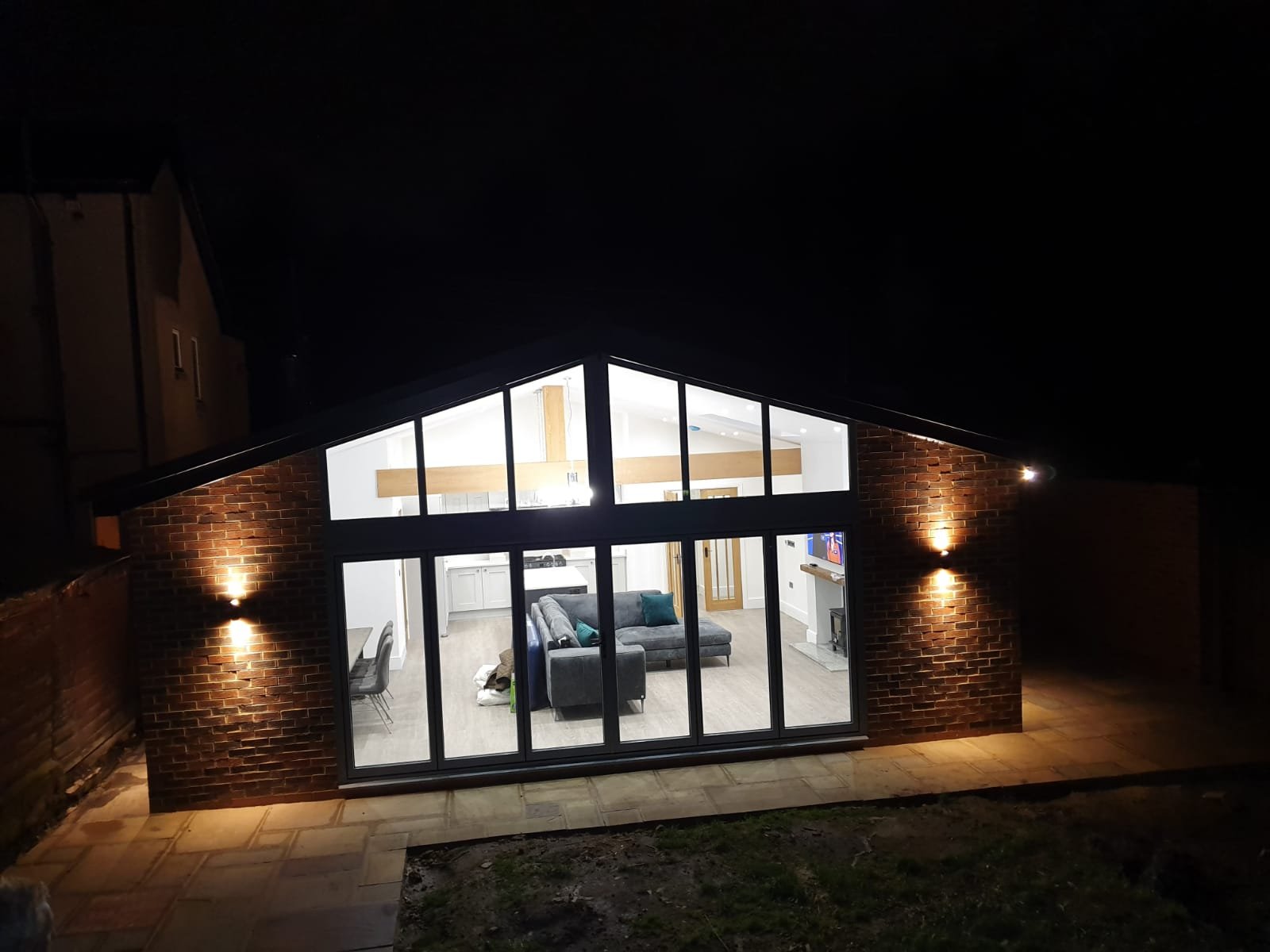 Nighttime view of a modern house with large glass windows revealing a brightly lit living room inside, brick exterior walls, and exterior wall-mounted lights.
