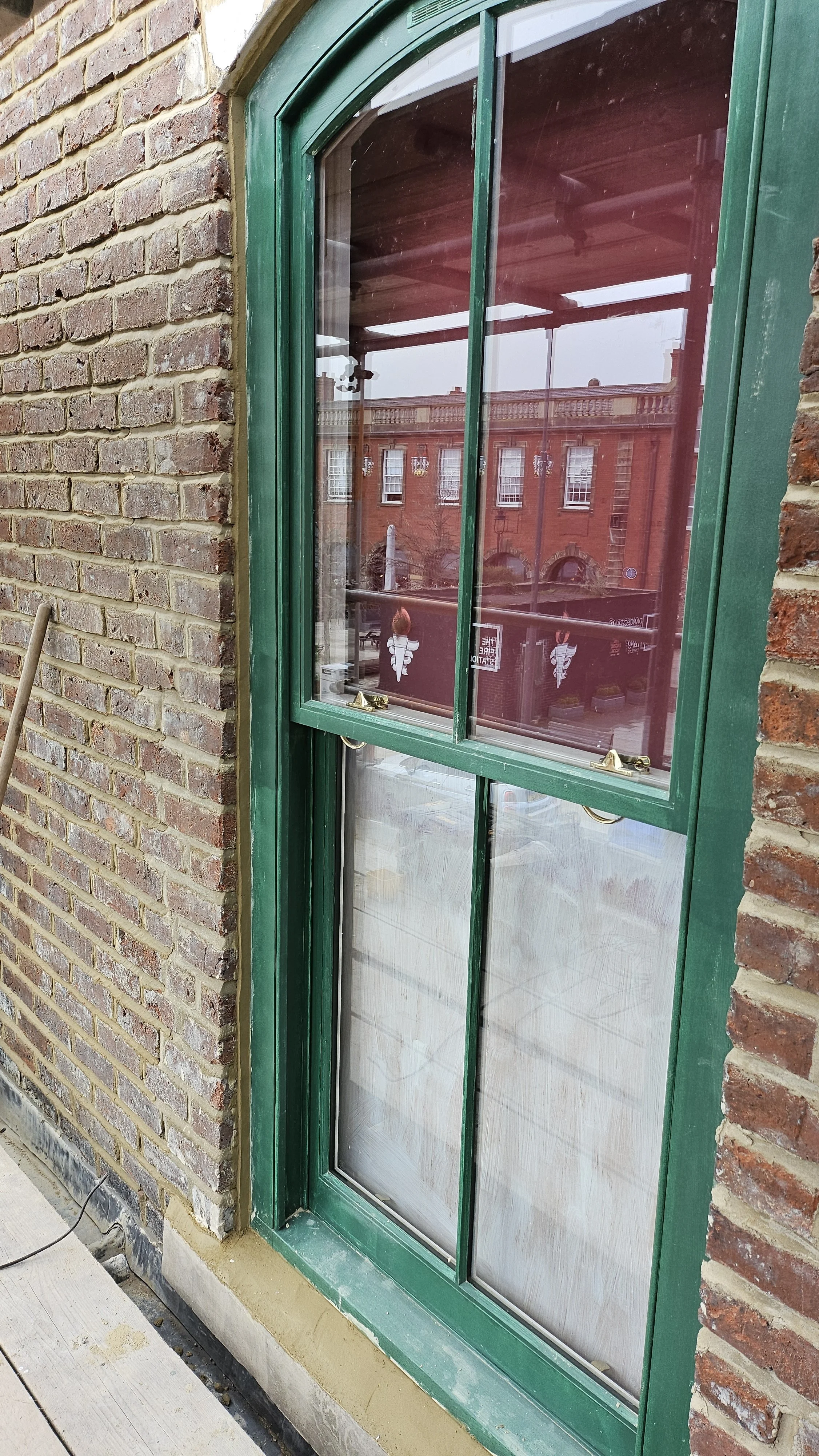 Close-up of a green-framed window with a brick wall to the left and an interior with red curtains. The window reflects a brick building across the street.
