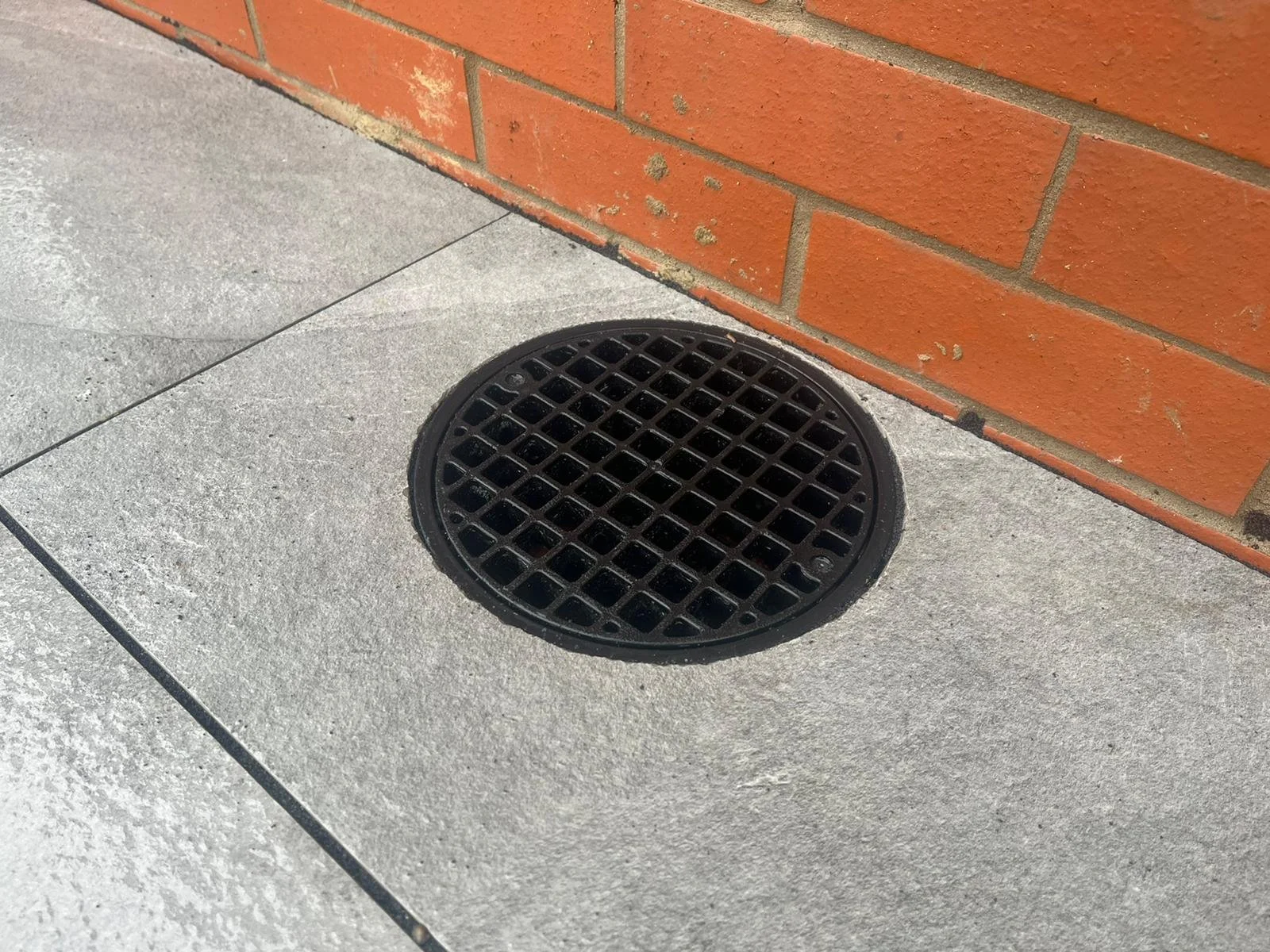Black round drain cover on concrete sidewalk next to a red brick wall.