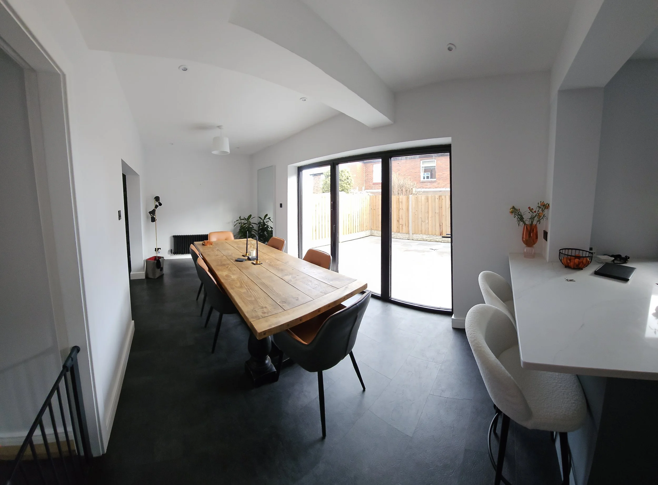 Modern dining room with a large wooden table, black and tan chairs, sliding glass doors leading to an outdoor patio, and a white countertop with bar seats, a vase with flowers, and a bowl of oranges.