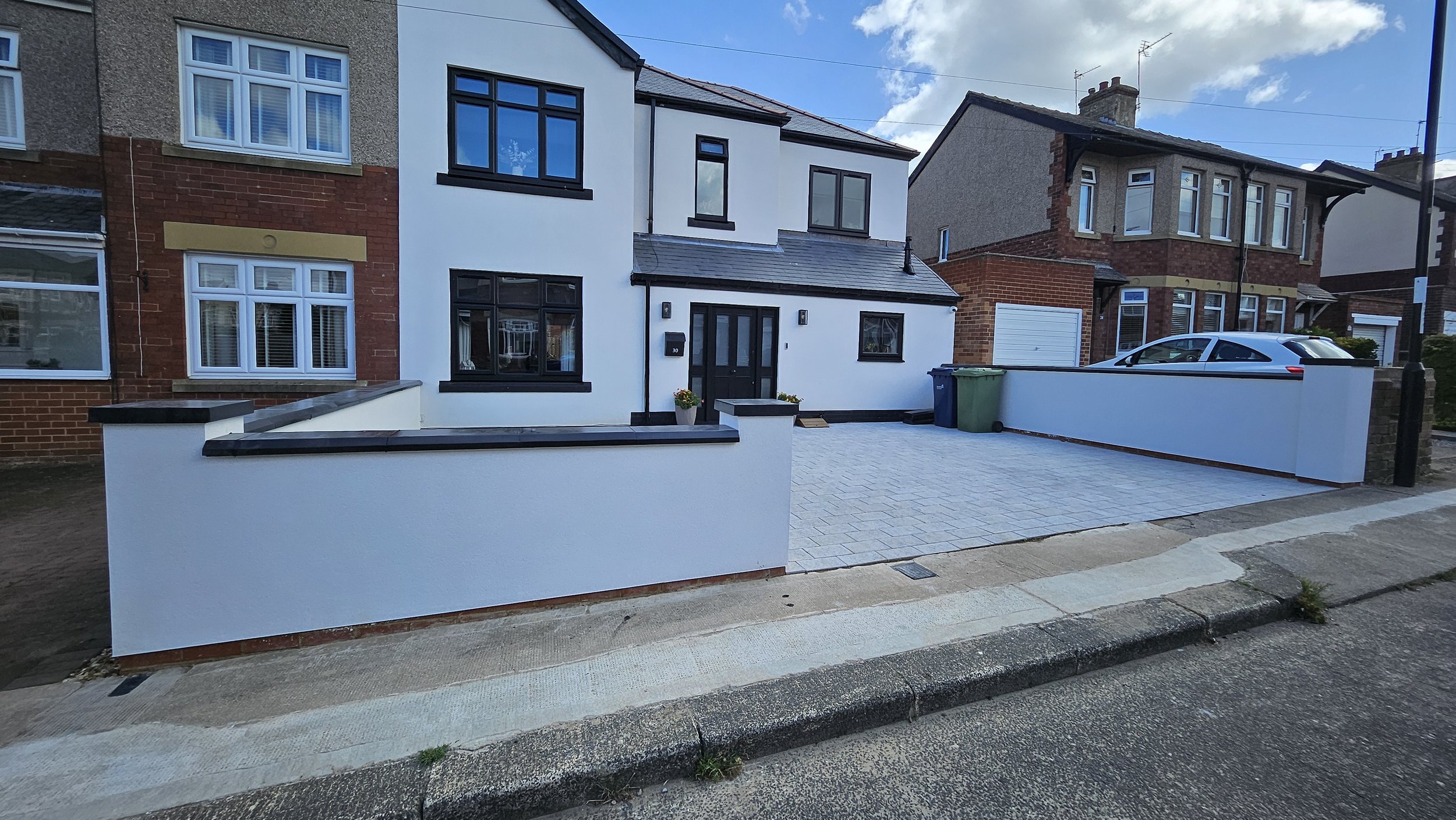 Front view of a modern white house with black window frames, a paved driveway, and a low white wall with black trim. There are two potted plants near the front door and a green trash bin on the driveway.