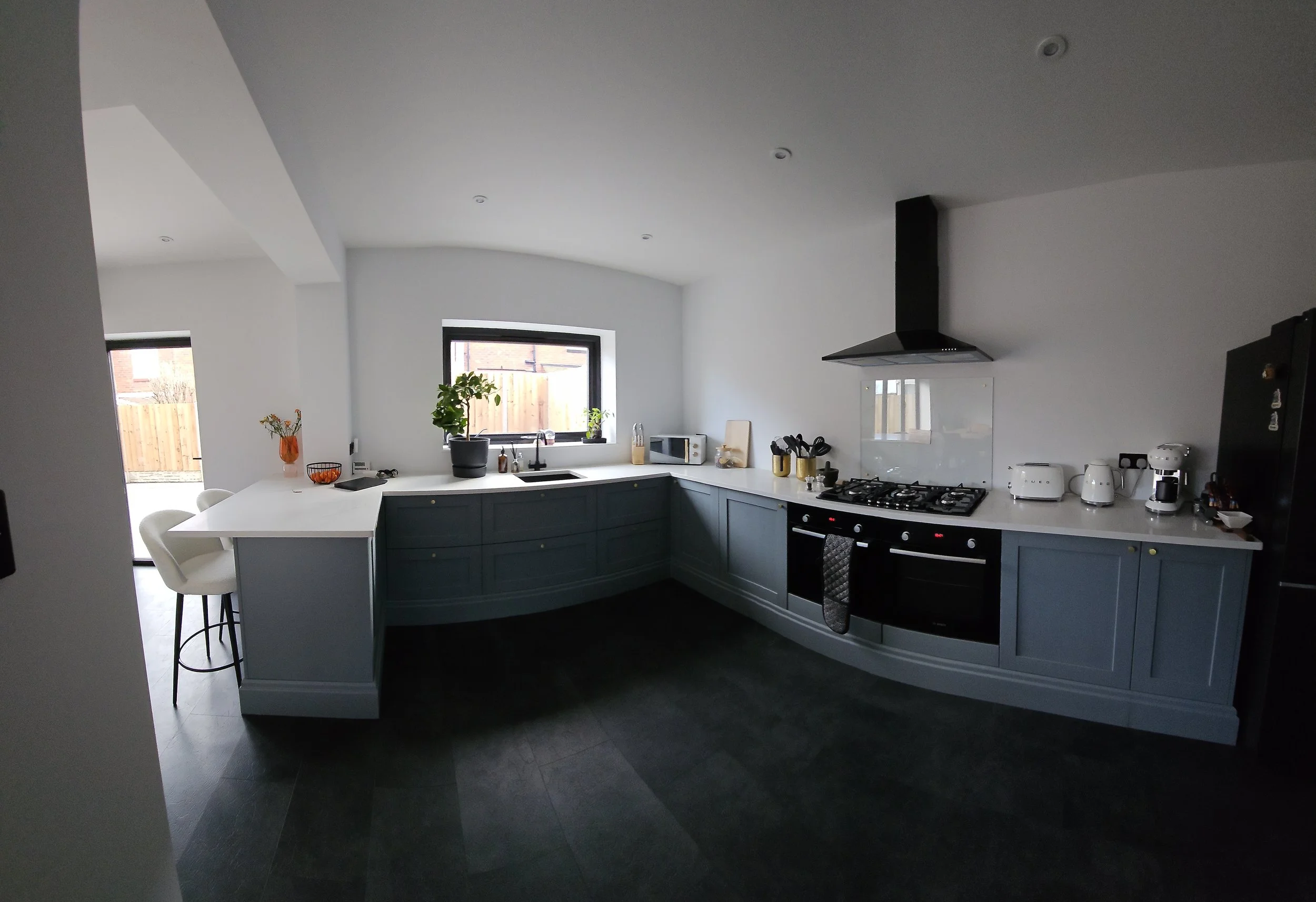 Modern kitchen with white and blue cabinets, black stove, and black range hood. Contains kitchen appliances and plants, with a window and small dining area.