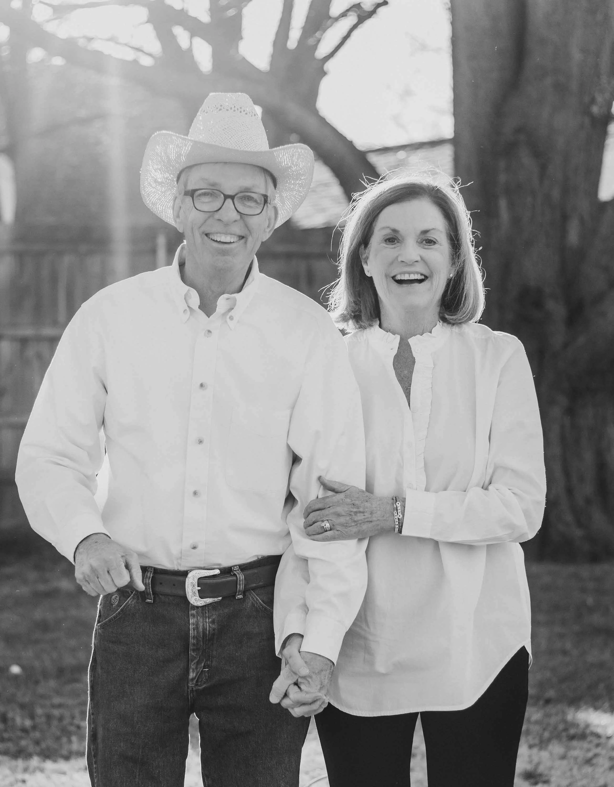 An elderly man and woman smiling and holding hands outdoors in a backyard with trees and a wooden fence. The man is wearing a cowboy hat, glasses, and a white shirt, while the woman is wearing a white blouse.