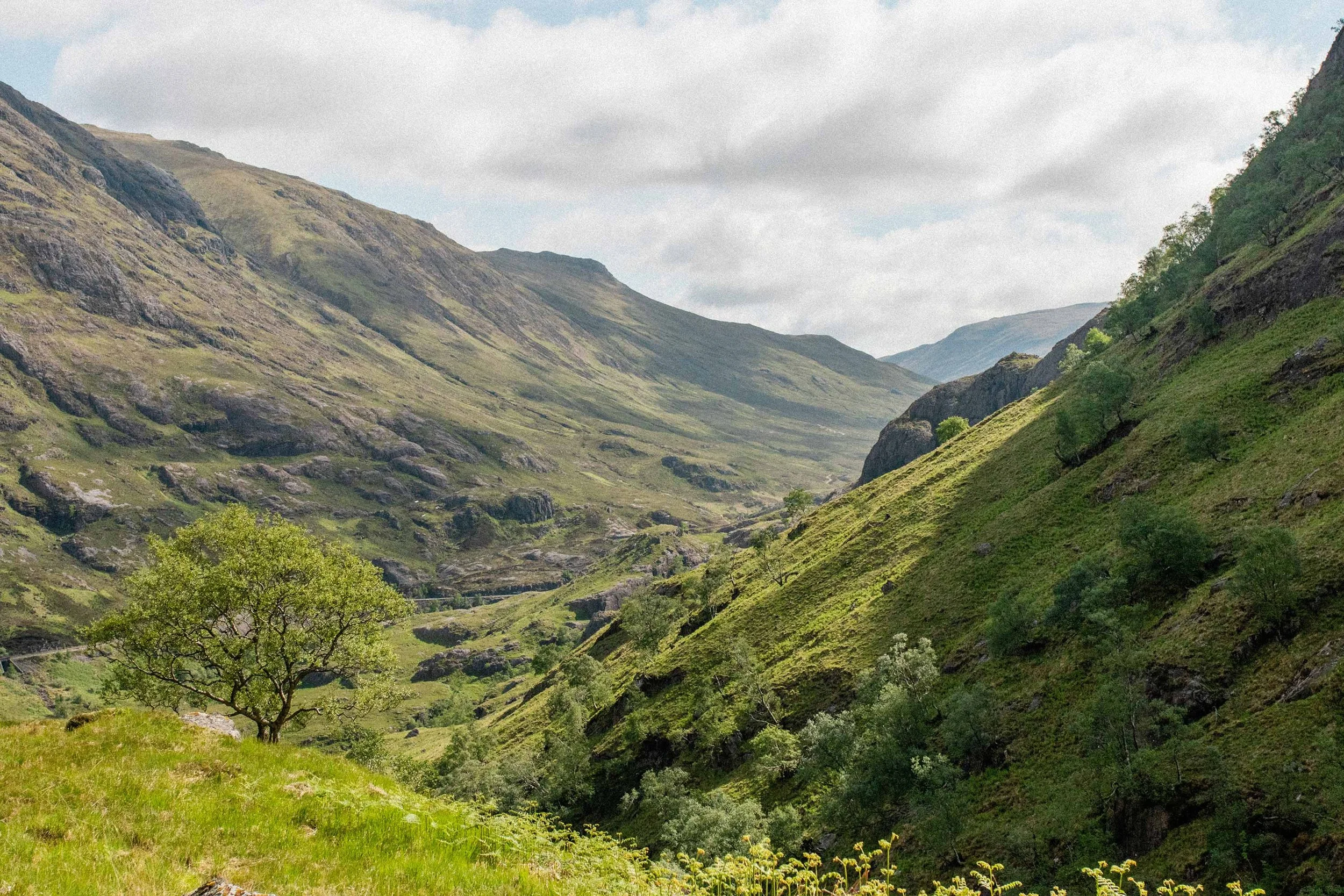 A lush green, Scottish valley surrounded by steep, rocky hills under a partly cloudy sky.
