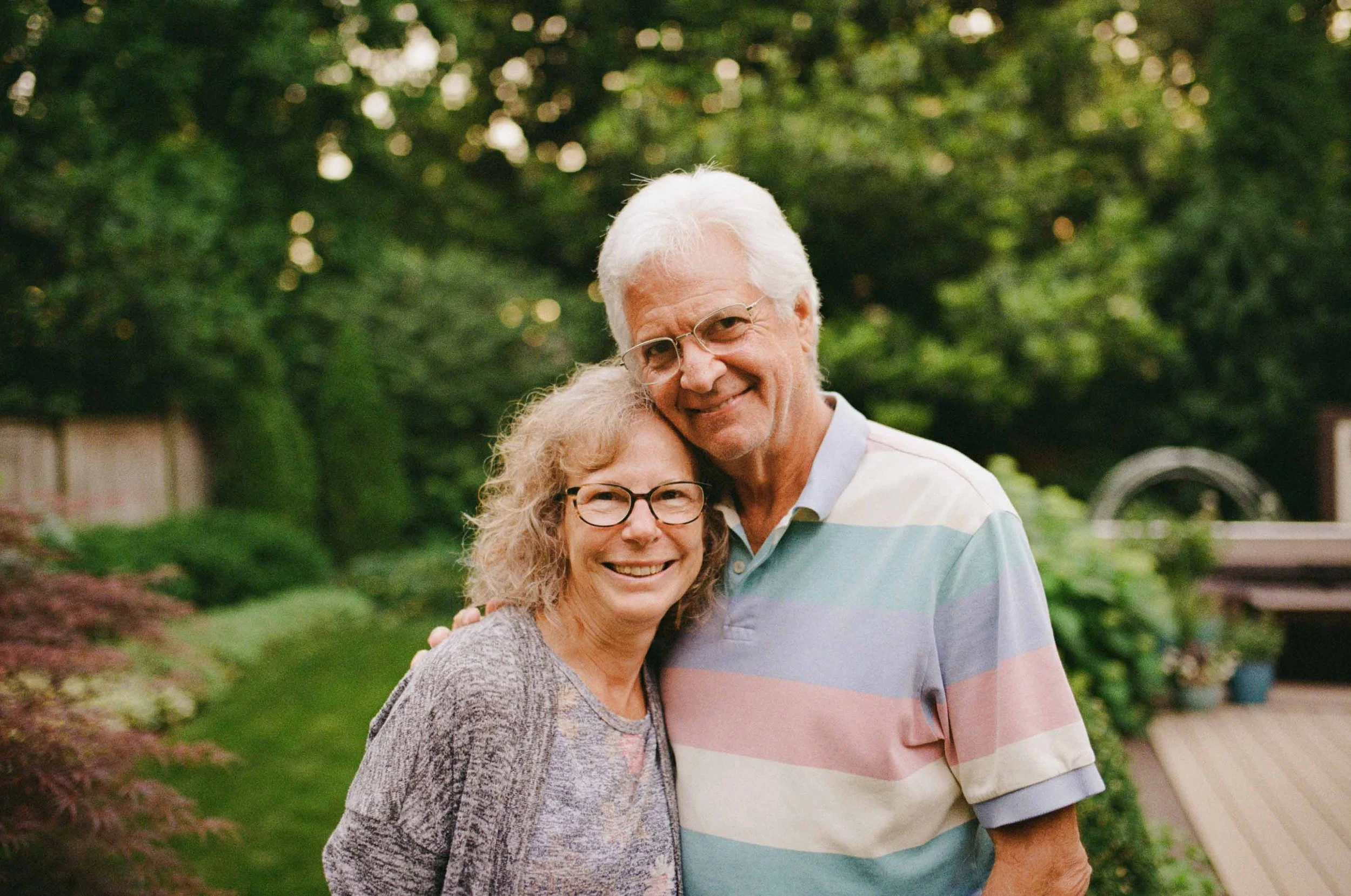 A smiling elderly man and woman hugging outdoors in a green garden.