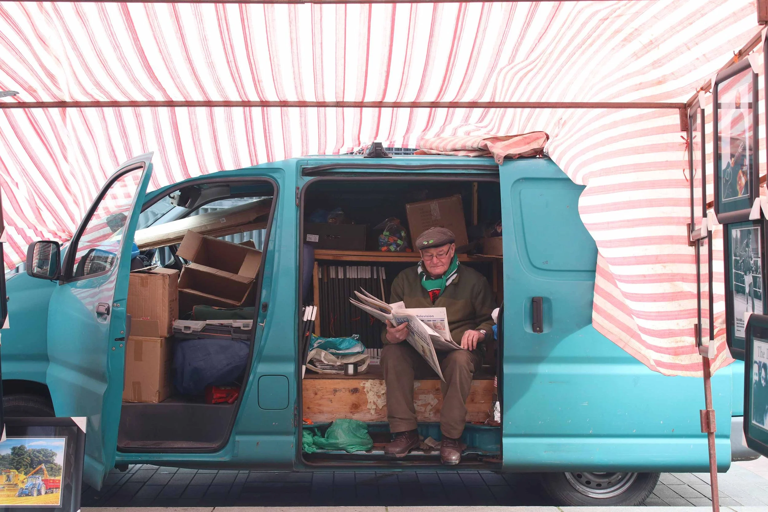 An older gentleman with glasses and a scarf sits in a turquoise van filled with boxes and documents, reading a newspaper under a red and white striped canopy at an outdoor market.