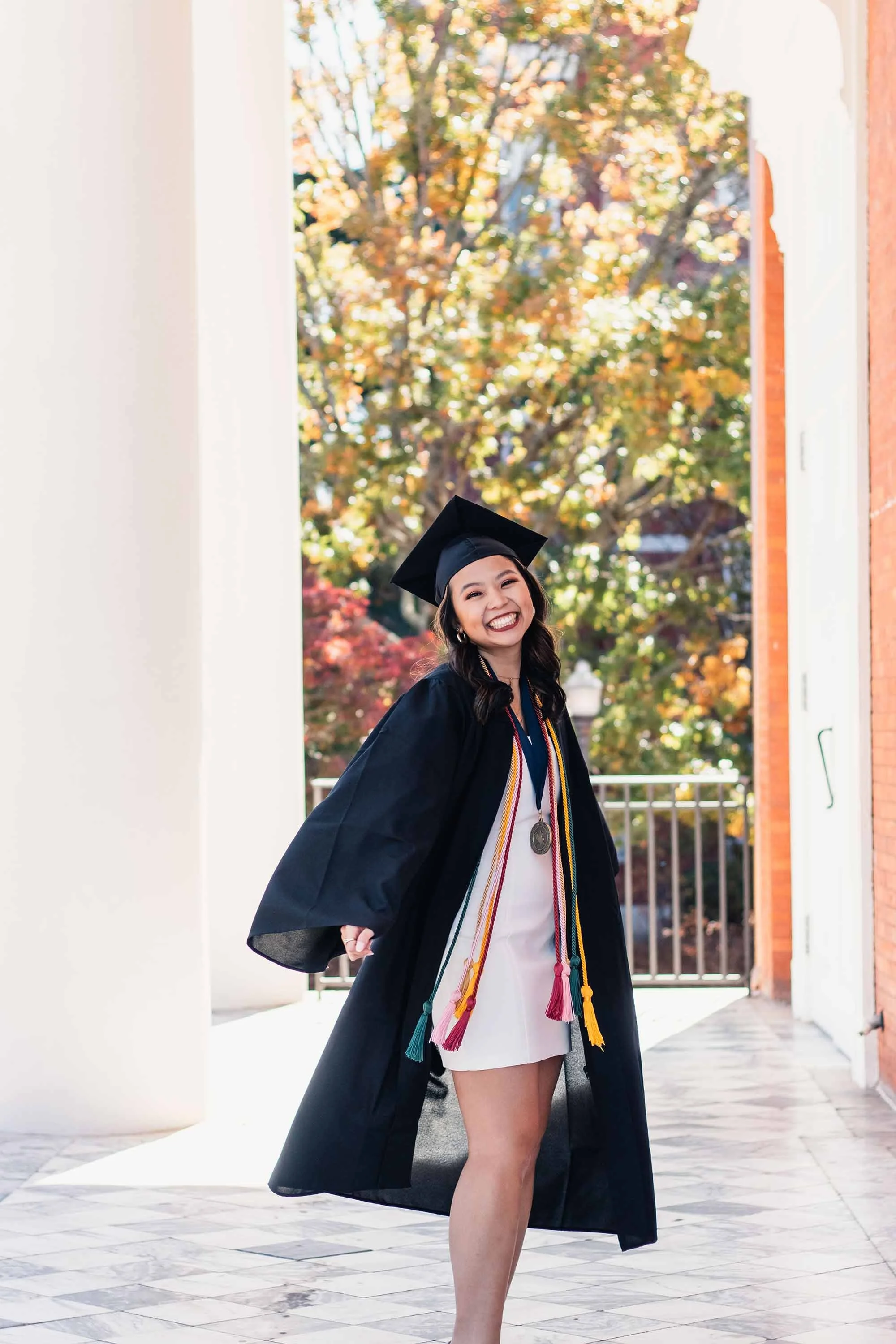 A young woman in a graduation cap and gown smiling outside on a bright fall day.