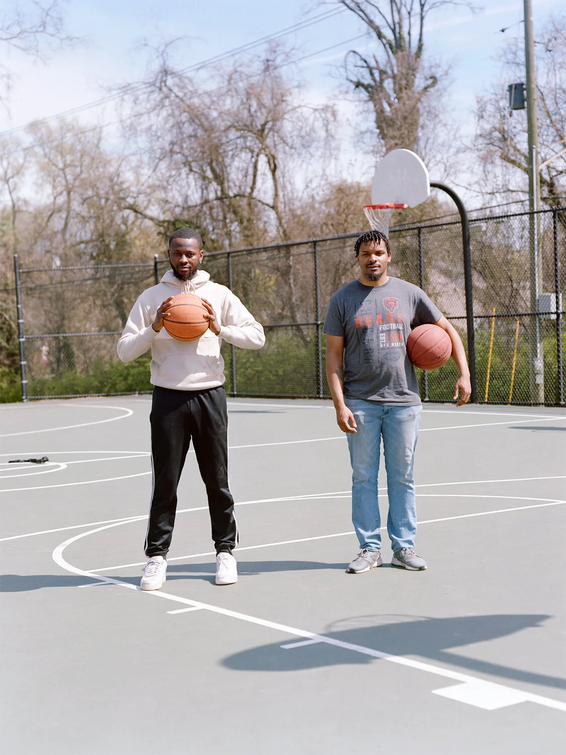 Two men standing on a basketball court holding basketballs, with a basketball hoop and backboard behind them.