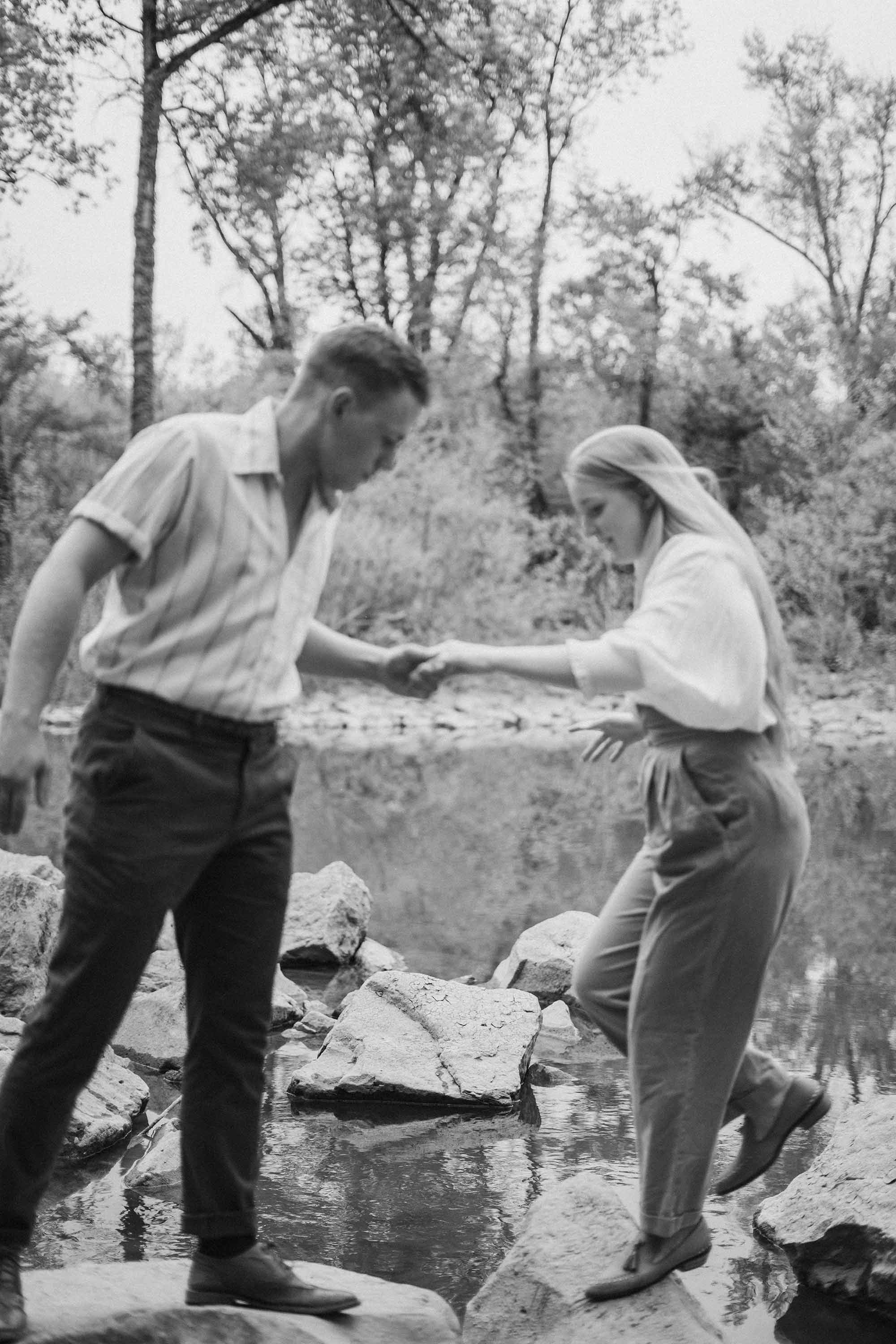 A black and white photo, showing motion, of a man and woman jumping from rock to rock in a creek, surrounded by trees. He offers a hand to support her.