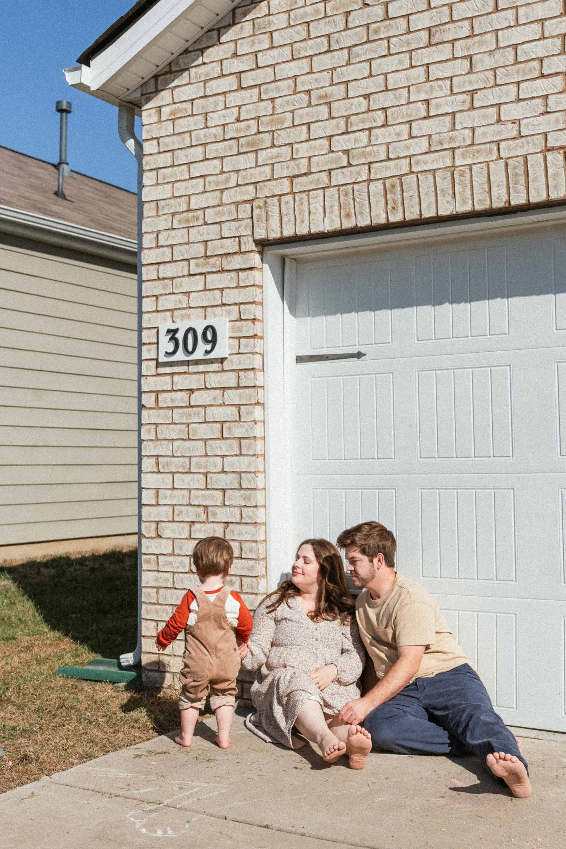 A family of three sitting on a sidewalk outside a house. The mom and dad sit close to each other, smiling, while their young child stands with his back to the camera, facing his mom.