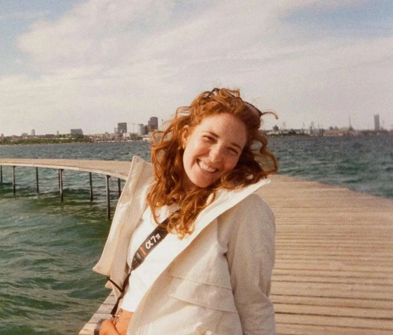 A woman with red curly hair smiling on a wooden pier by the water with a city skyline in the background.