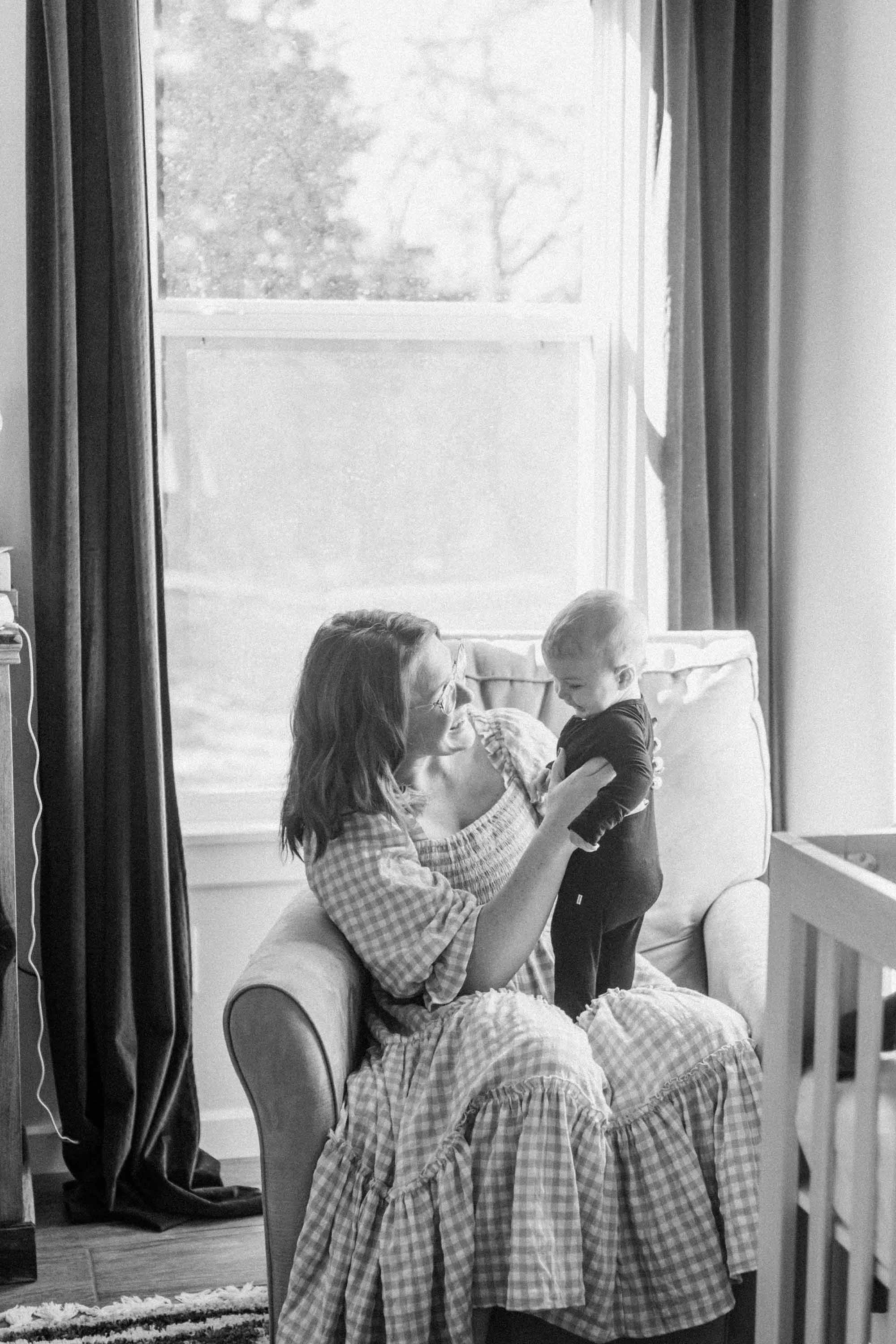 In black and white, a mom, in a checkered dress, sits in an armchair near the window holding her baby.