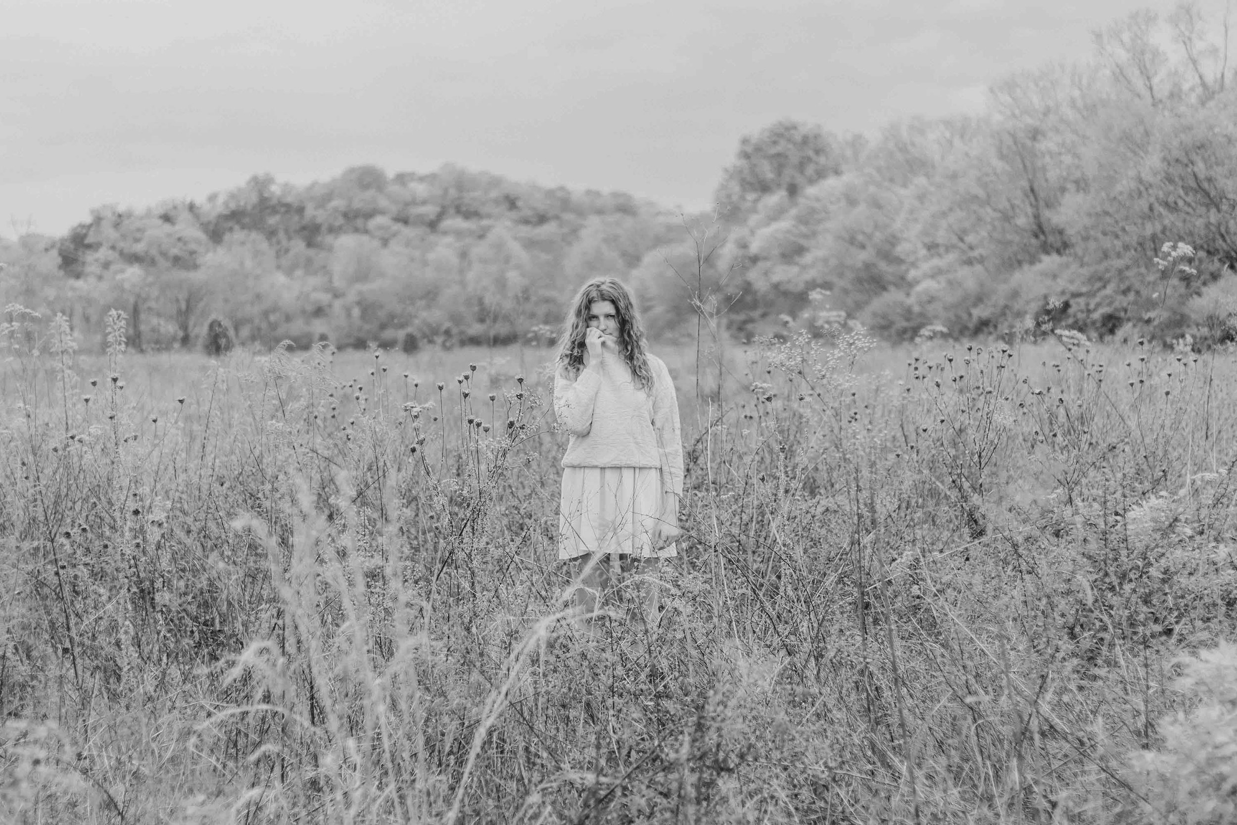 A black and white photo of a young girl standing in a field of tall grass and wildflowers, with trees and hills in the background.