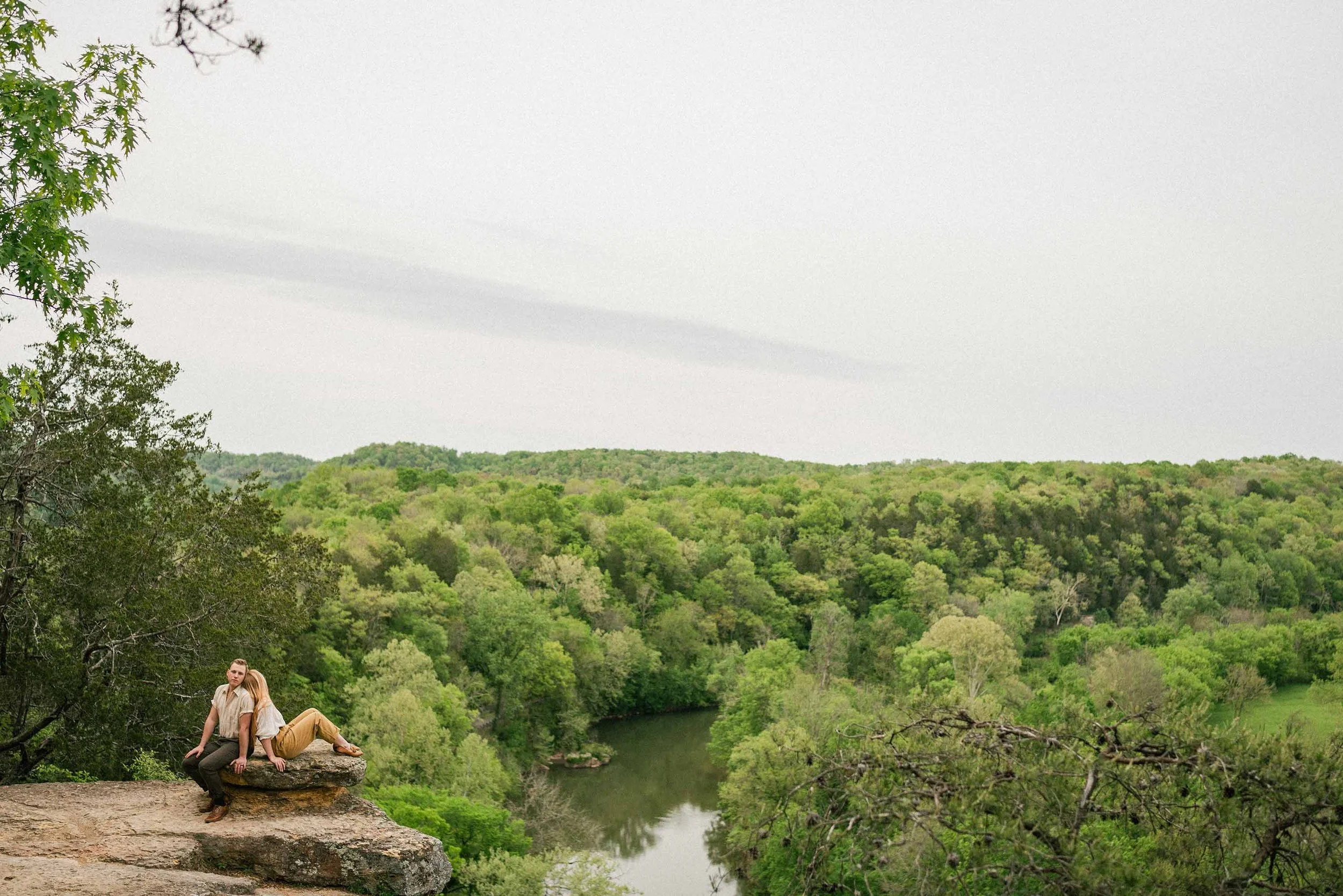 A young couple, a man and a woman, sit back-to-back on a large rock overlooking a lush green valley with a river below, under a cloudy sky. He looks at the camera, she looks at the view out and below.