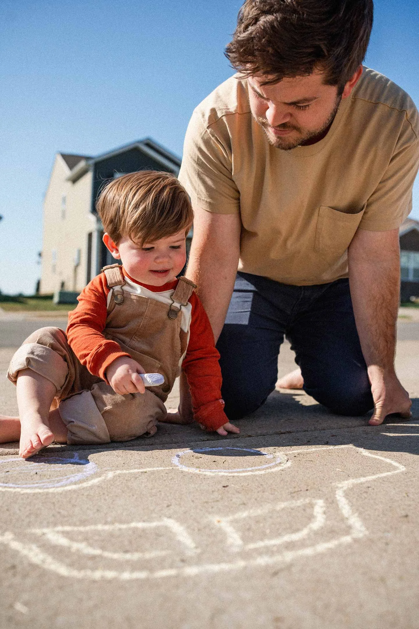 A young child and his father, sit on the sidewalk playing with chalk on a sunny day in the neighborhood.