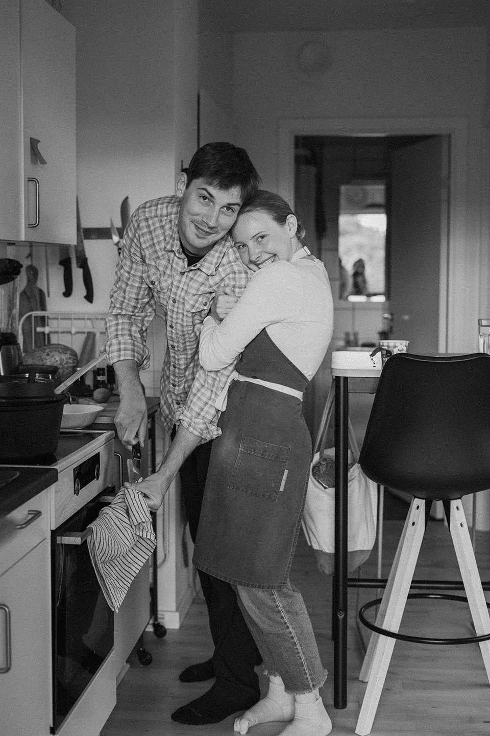 A couple enjoys a moment in the kitchen, with the man leaning over the stove and the woman embracing him, both smiling.