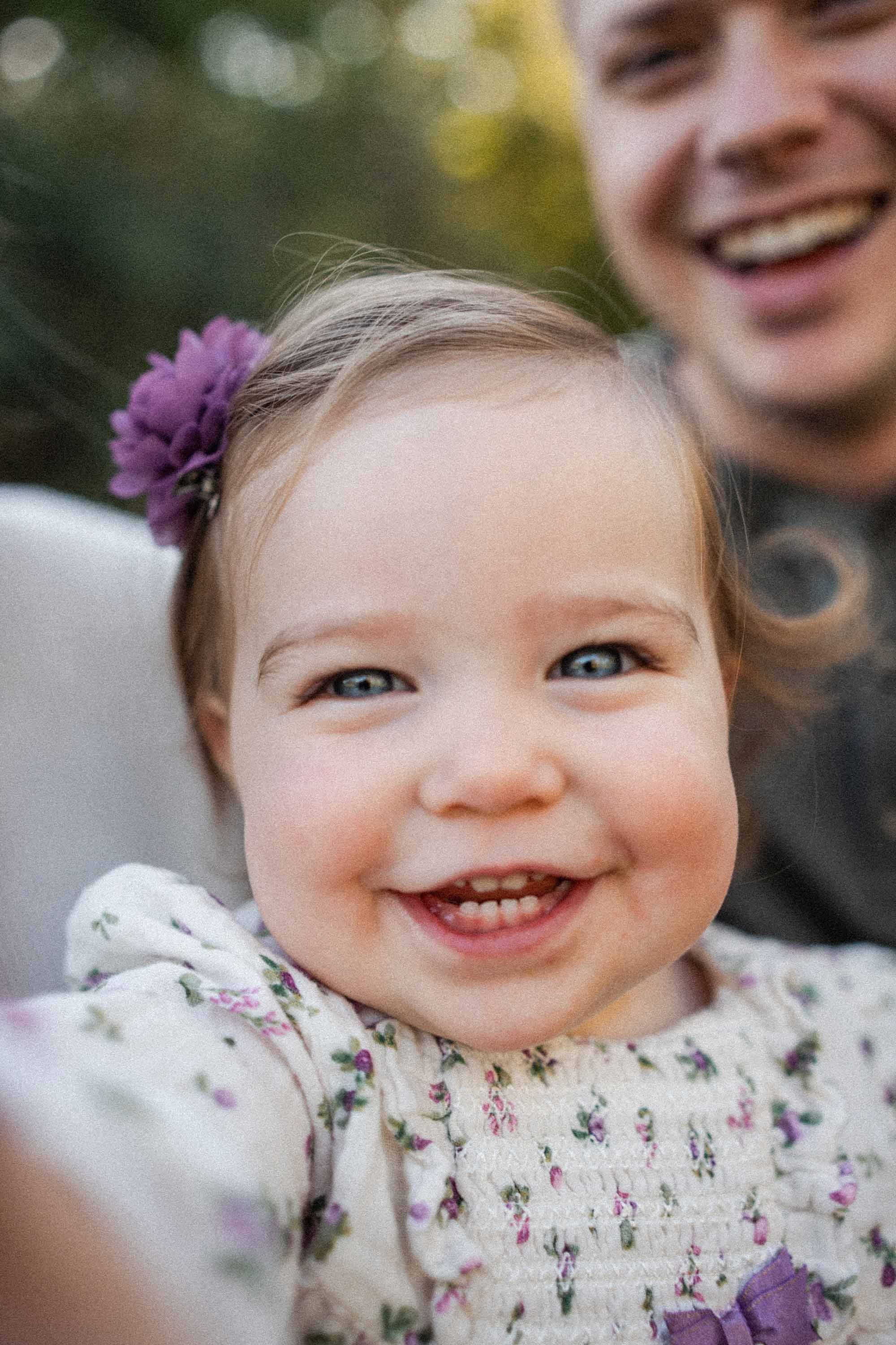 Close-up of a smiling young girl with blue eyes, wearing a floral dress and a purple flower in her hair, reaching for the camera, with her father smiling in the background.