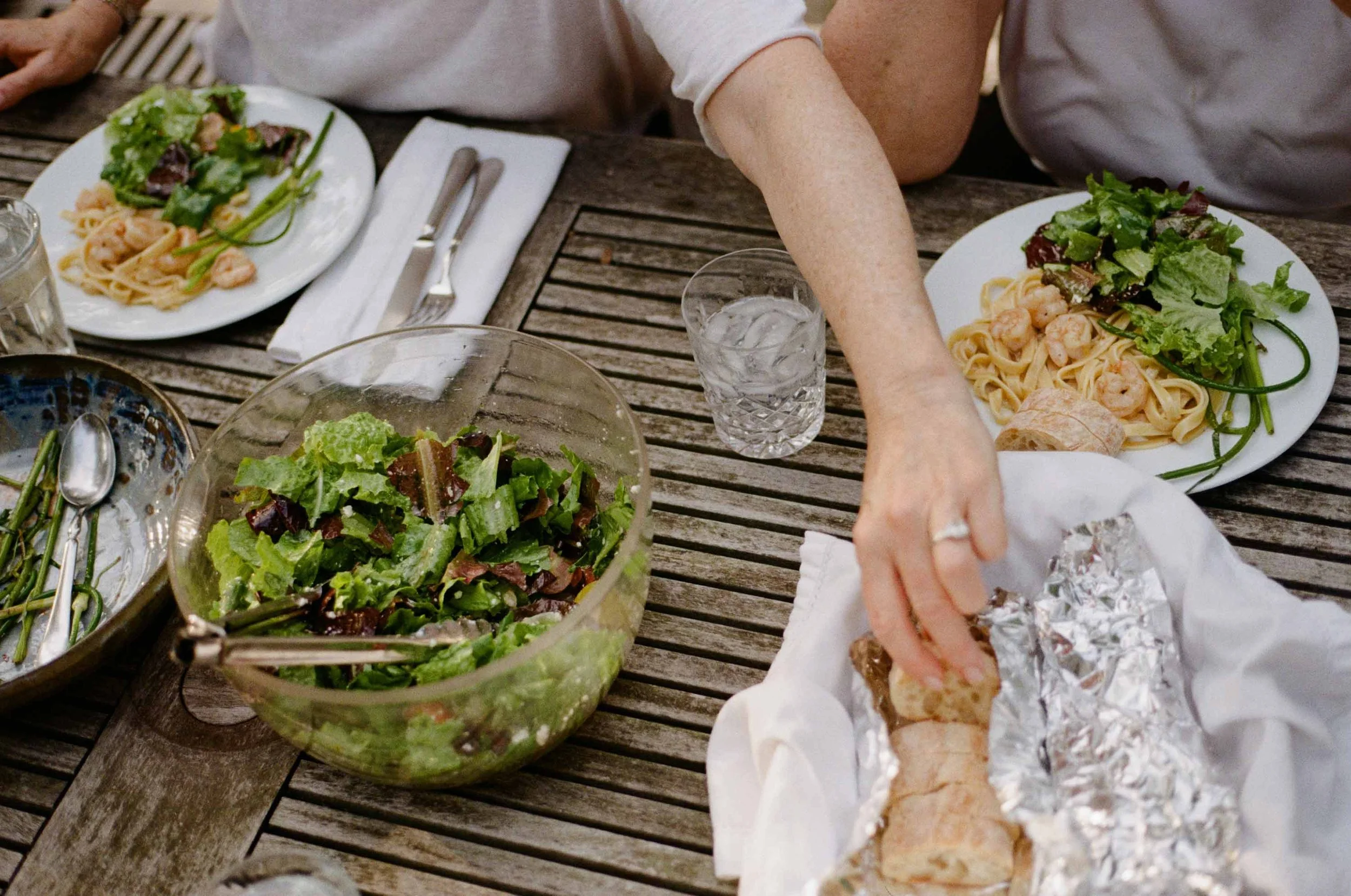A color film photo of a outdoor dinner table with fresh farm made dinner including bread, salad, and pasta on an outdoor table. A woman reaches for the bread.