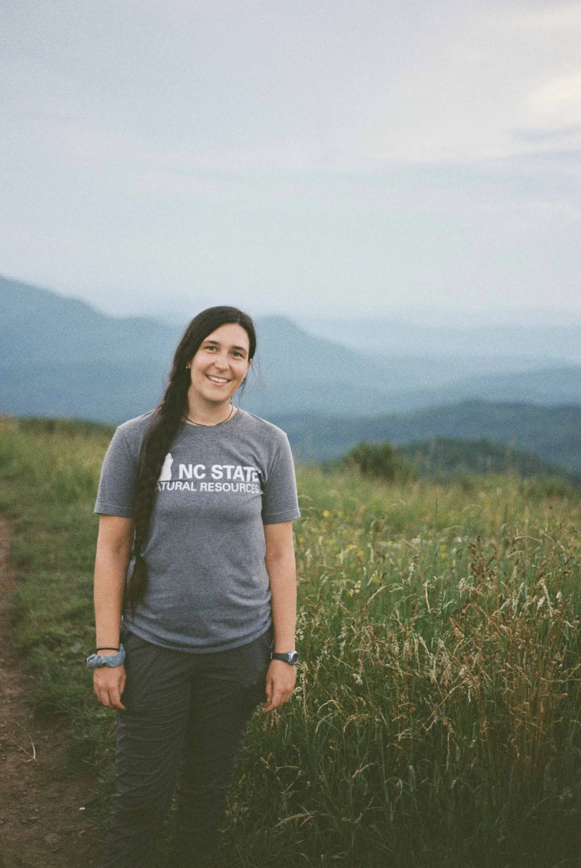 A color film photo of a young woman standing on a grassy trail in the Smokey Mountains, smiling, wearing a gray NC State Natural Resources t-shirt with a mountain range in the background.