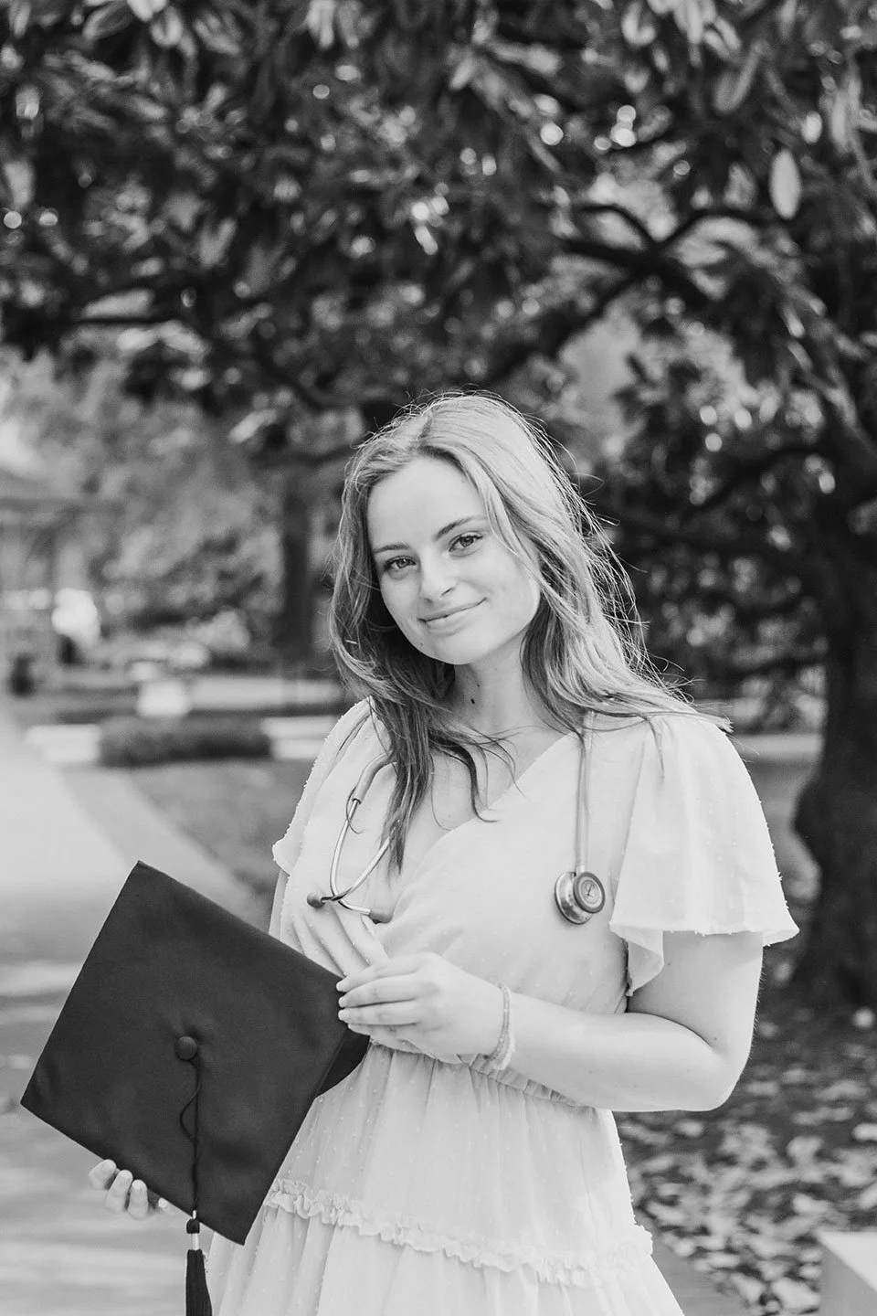 Black and white photo of a young woman in a light-colored dress holding a graduation cap, wearing a stethoscope around her neck, standing outside with trees in the background.