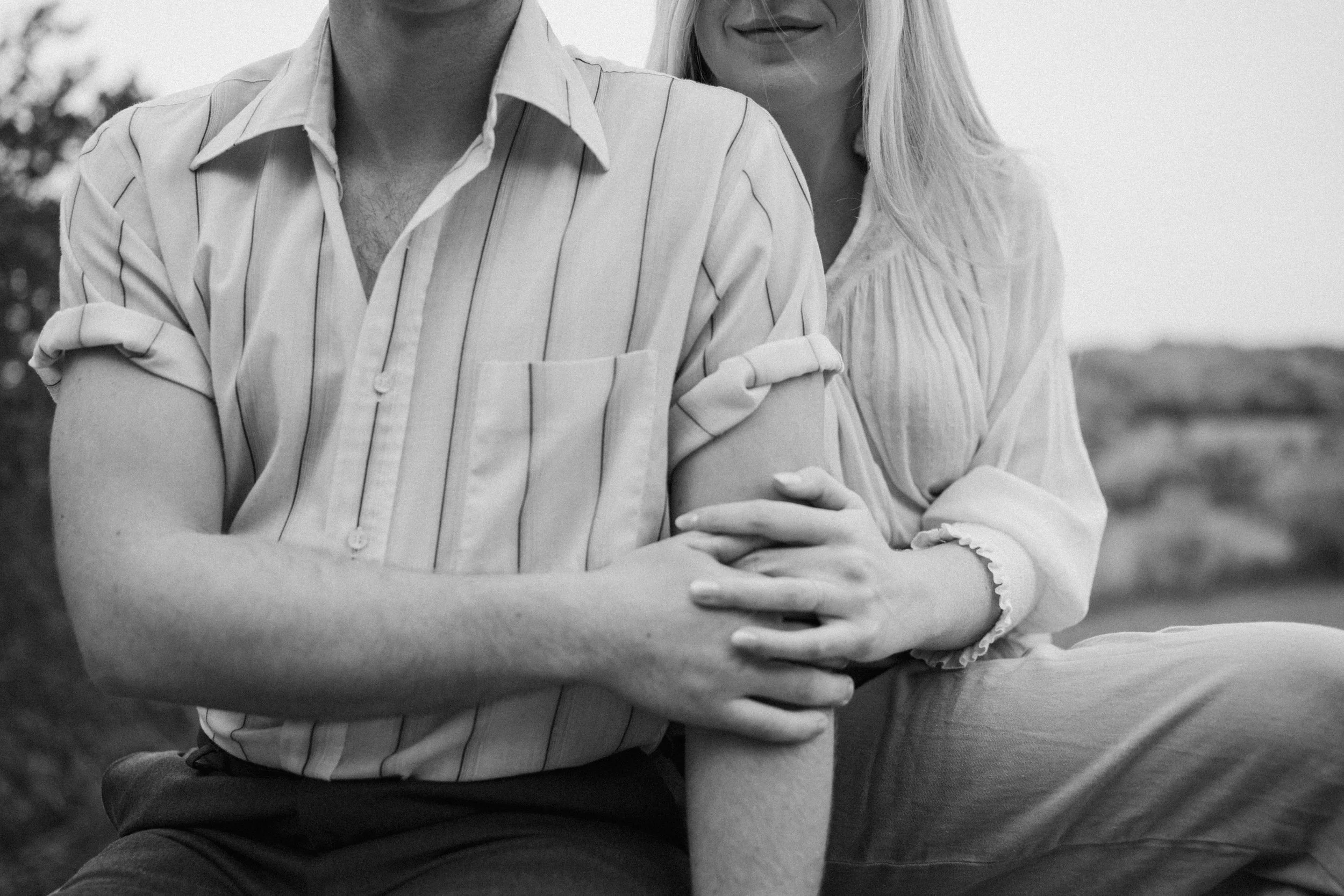 A black and white photo of a couple sitting in nature, viewed waist up to just under her nose. The woman sits behind the man, holding his arm with her hands as he reaches over to hold her hand.