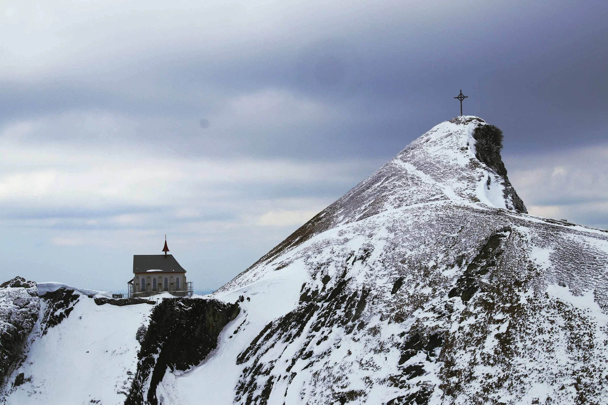 Snow-covered mountain with a cross at the peak and a small chapel on a flat area to the left.