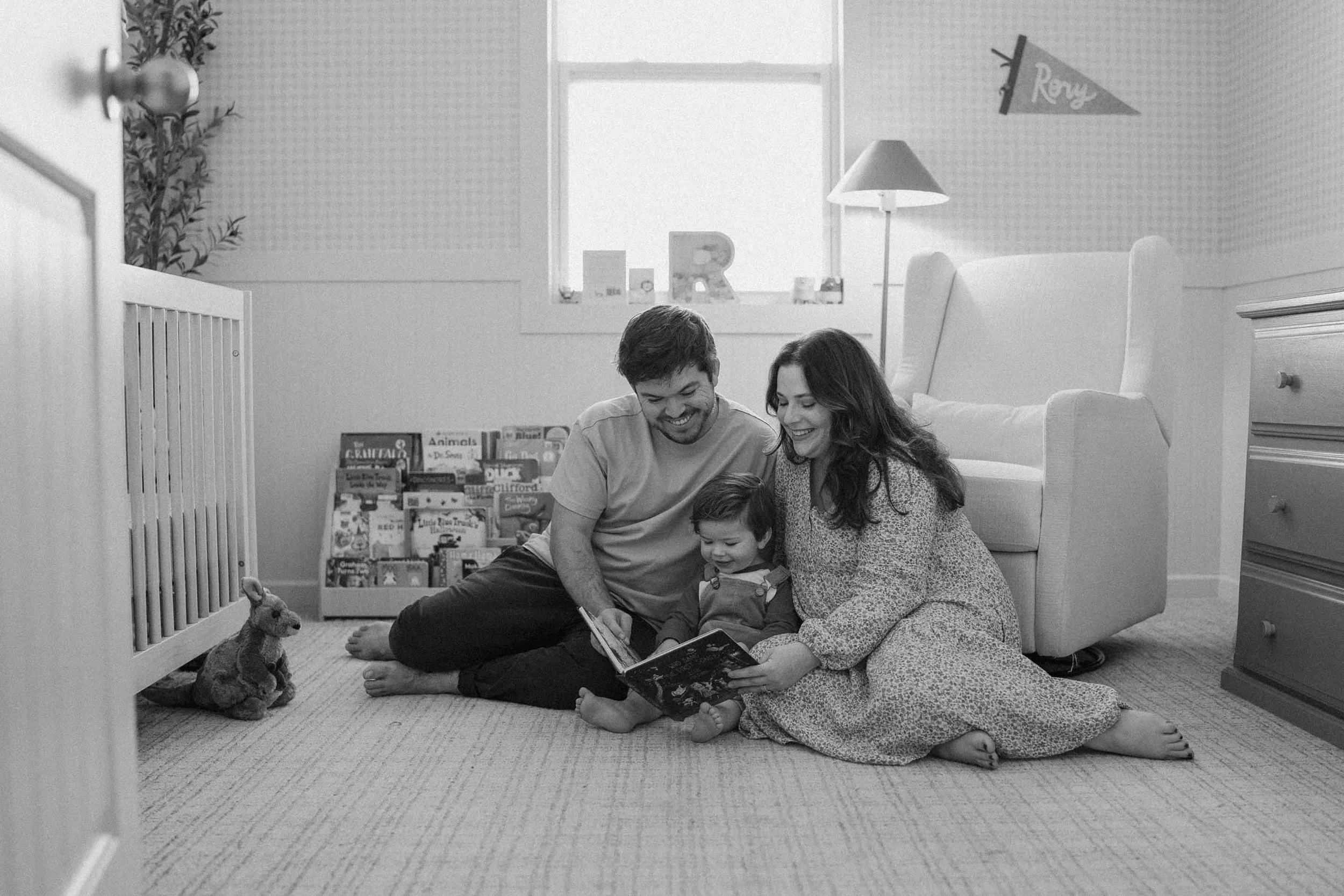 In black and white, a family of three sitting on the floor in a bedroom, reading a book together, with a stuffed animal and a bookshelf nearby.