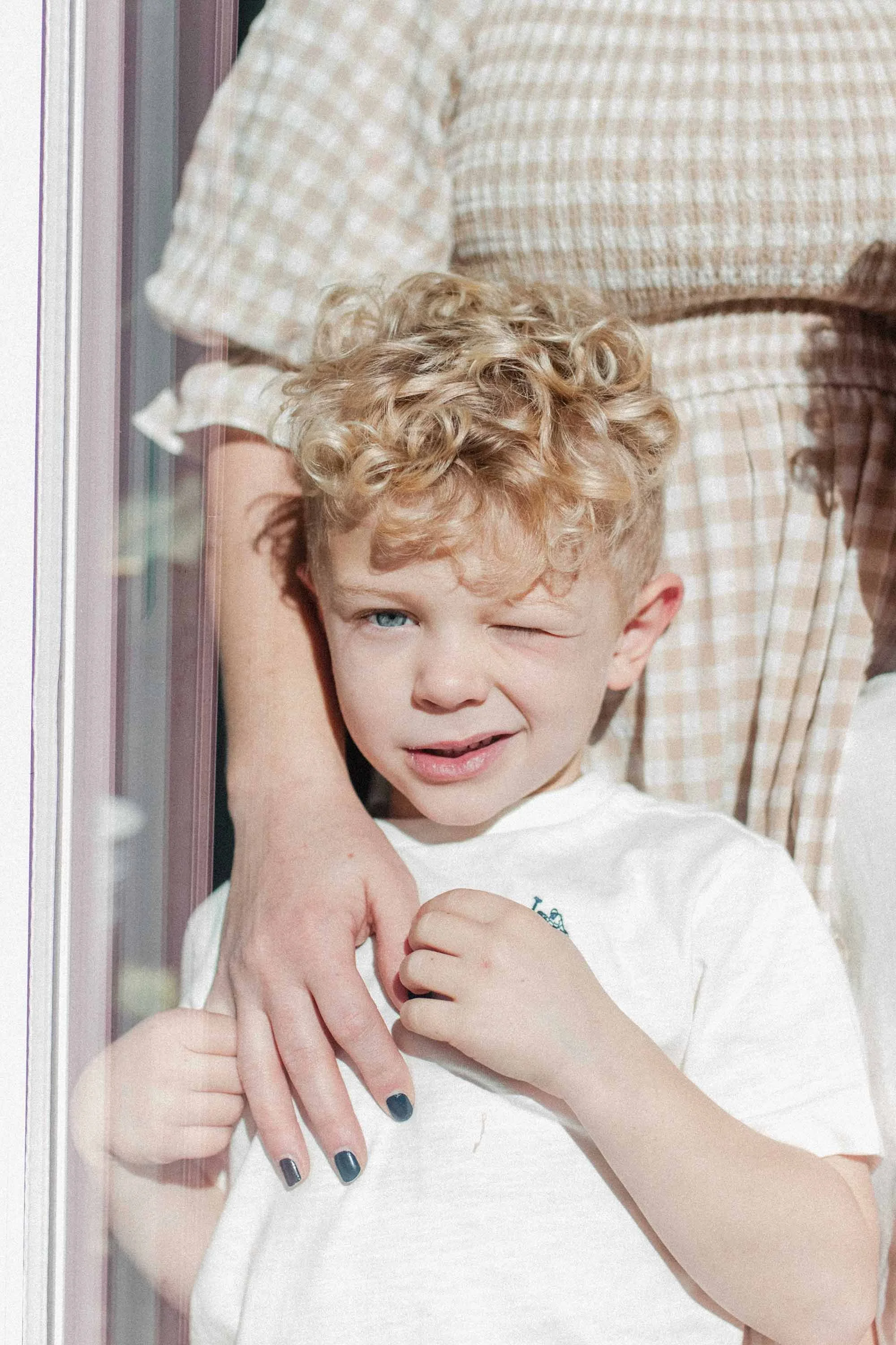 Through a window, young boy with curly blonde hair stands squinting in the sun while holding his mom's hand over his chest.