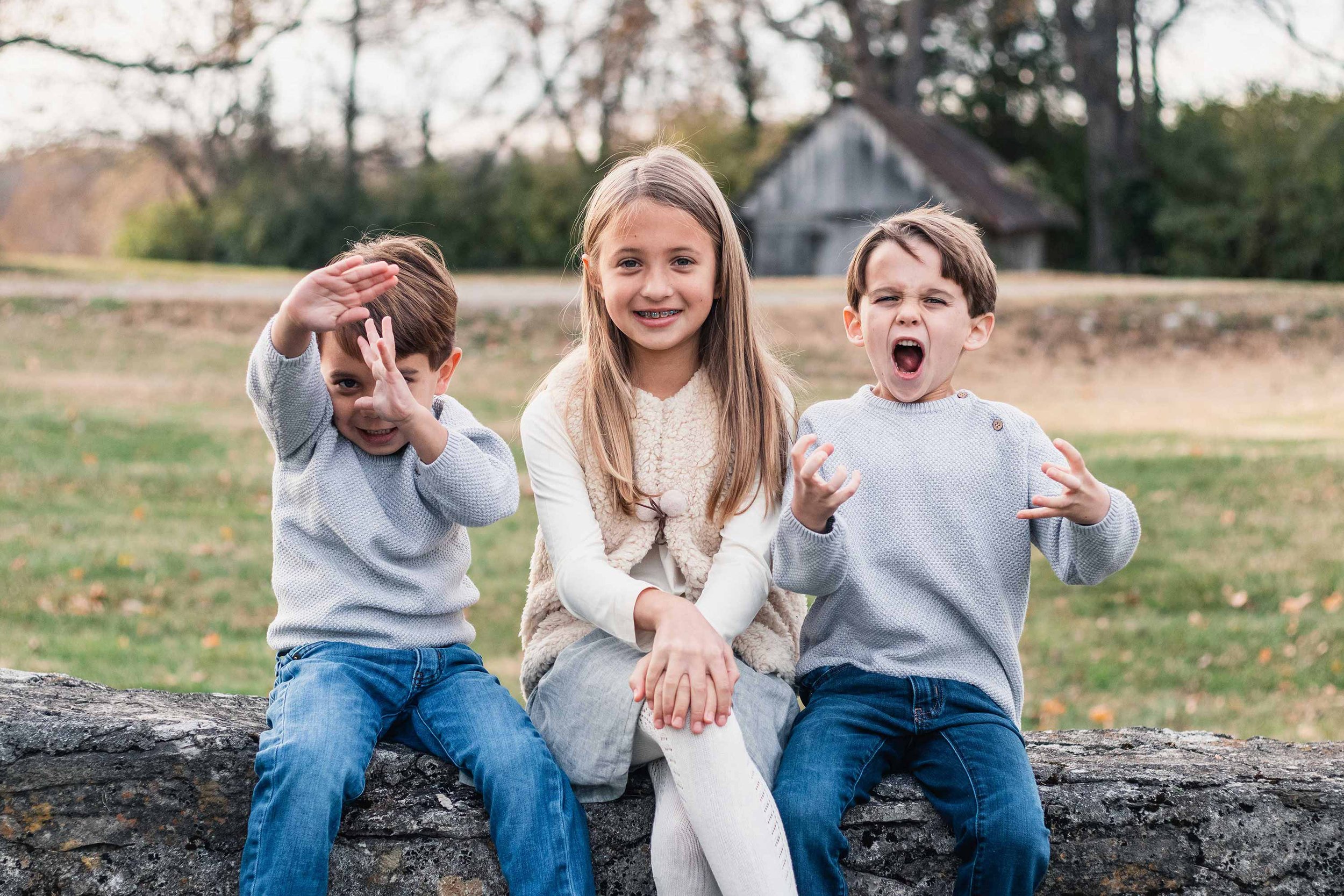 Three children sit on a wall outdoors showing unique and expressive faces. The boy on the left shows his ninja moves, the girl in the middle politely smiles, and the boy on the right with his mouth open and fingers curled in an excited expression.