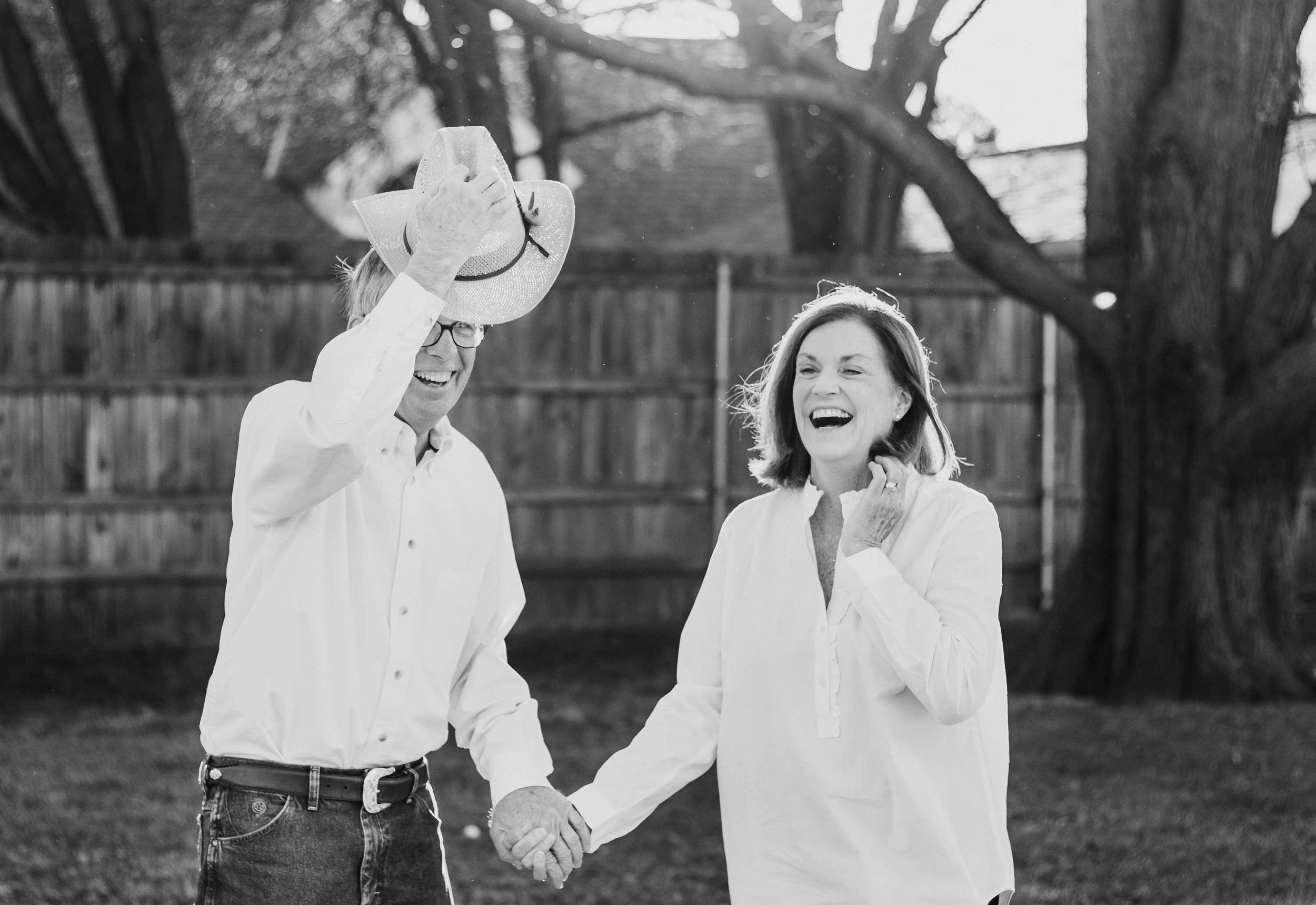 A happy older couple outside in a backyard, holding hands and laughing. The man tips his cowboy hat off his head, while the woman laughs.