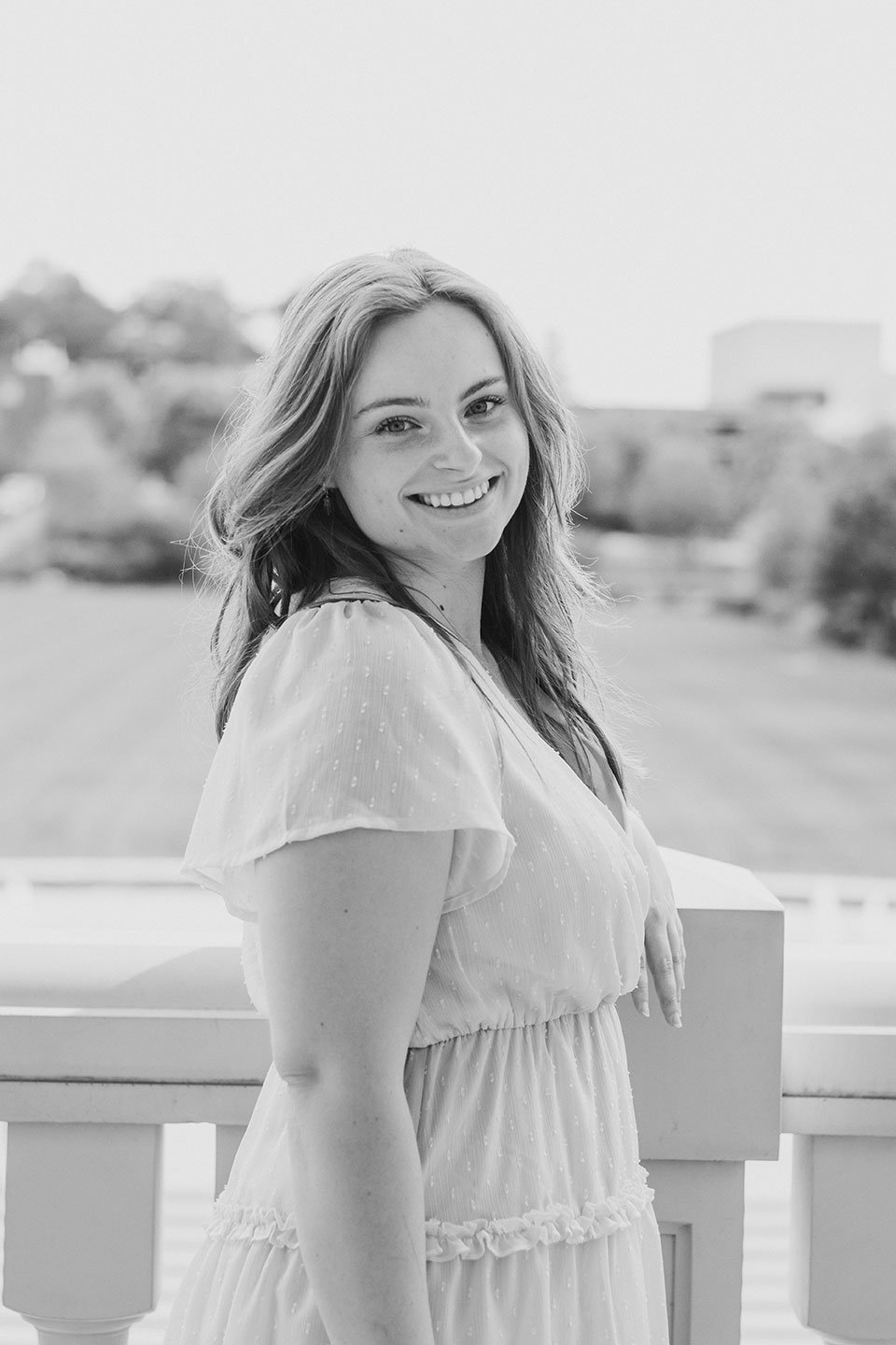 Black and white photo of a smiling young woman in a light dress with puffed sleeves standing outdoors with a blurred background of trees and buildings.