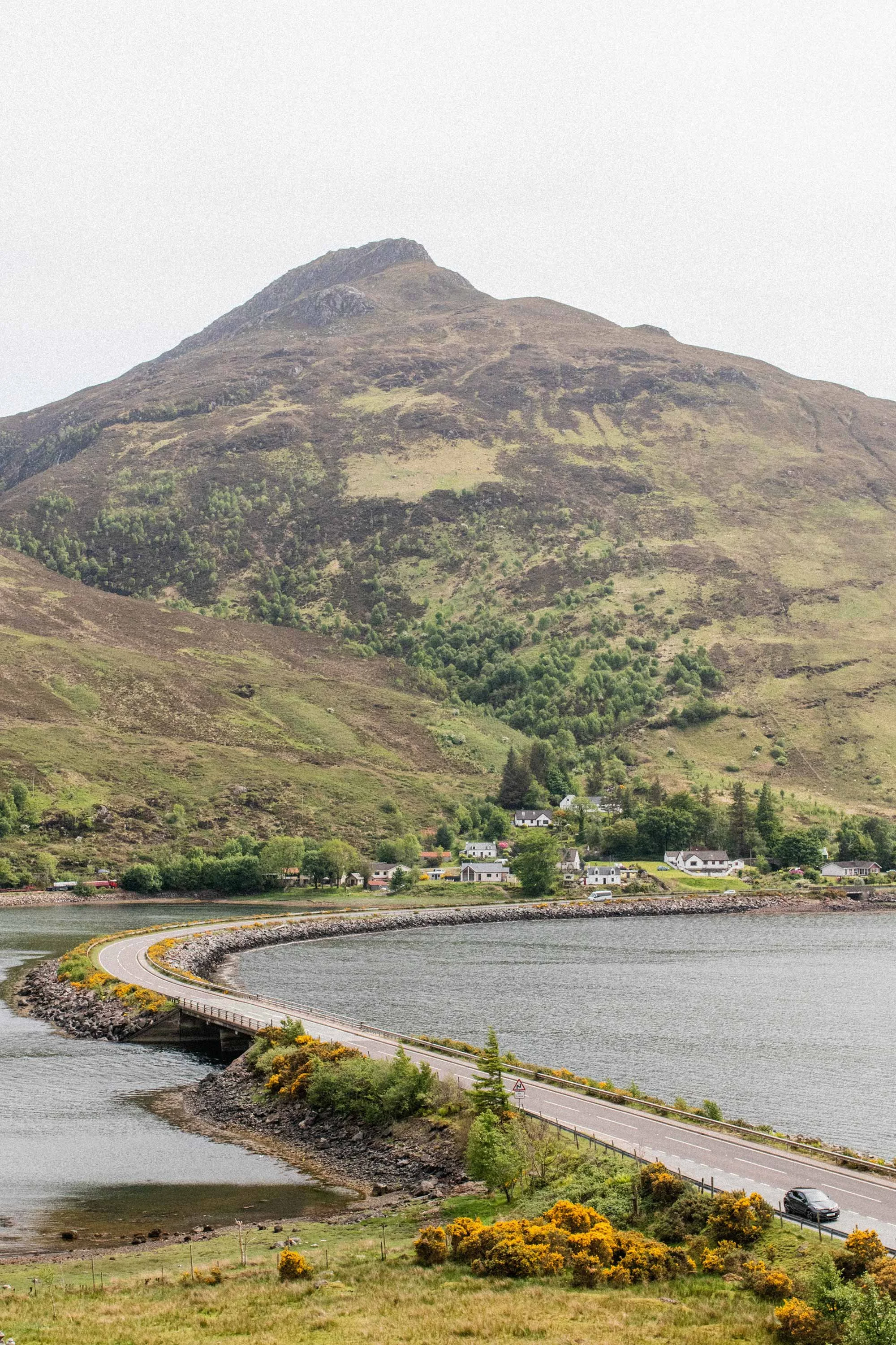 A mountain with a winding road along a body of water, and a small village with houses at the mountain's base in Scotland.