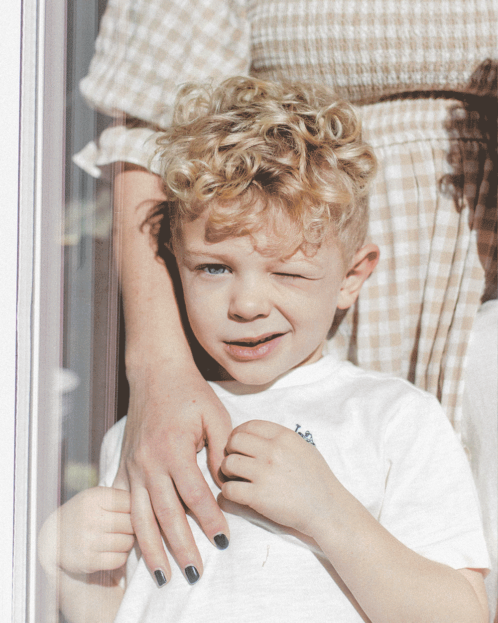 A young boy with curly blond hair winking and grimacing while being comforted by an adult, who is holding his shoulder outside a glass door.