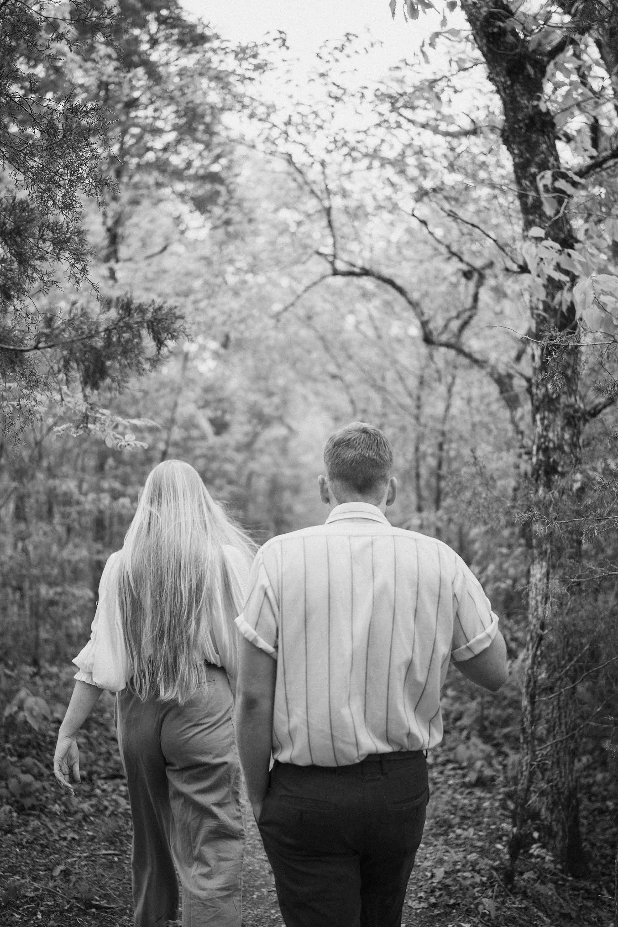 A black and white photo, showing motion, of a couple, man and woman, walking through a wooded forest, viewed from behind.
